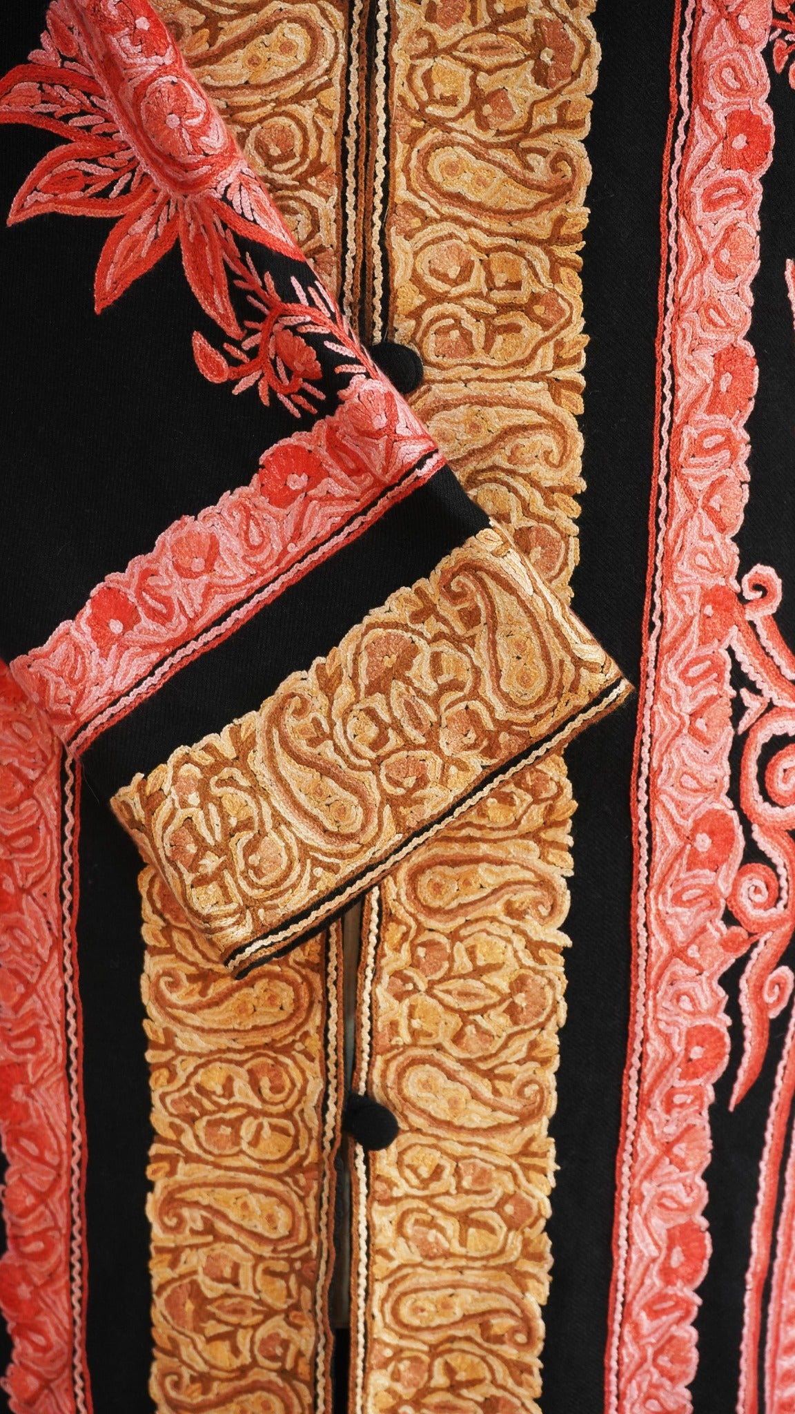 Detail of golden Aari paisleys on the black jacket’s crossed cuff beside coral-pink embroidered flowers and scrolls