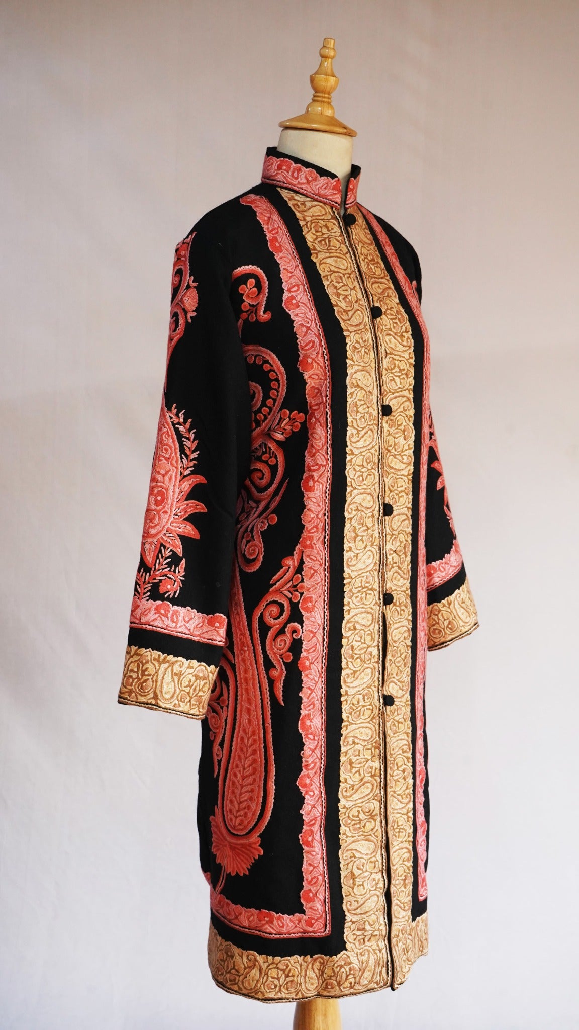 Angled full view of a black long jacket with golden front and cuff borders and sweeping coral-pink paisley embroidery