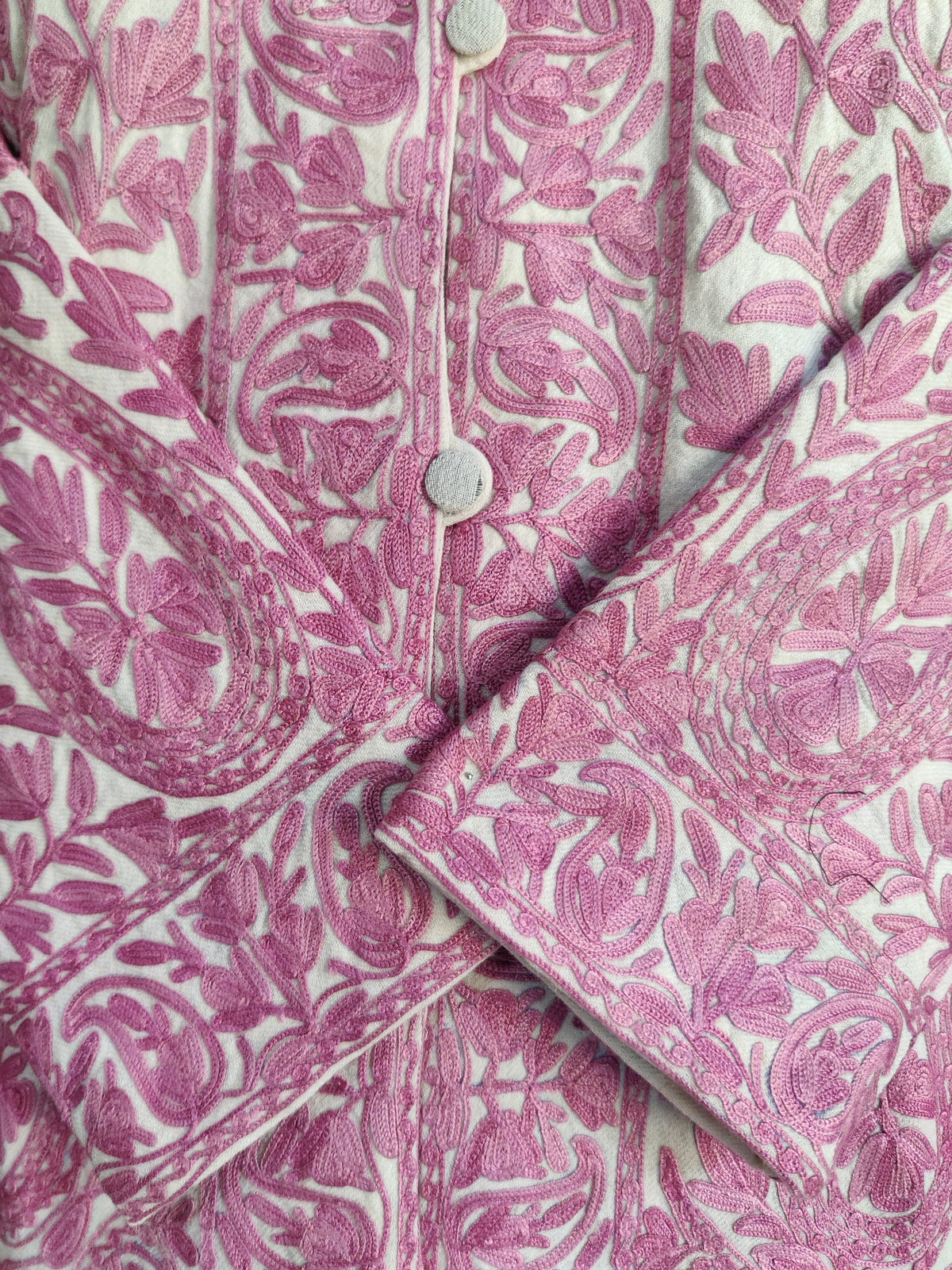 Close-up of raised pink Aari flowers and leaves on a cream jacket, with pale buttons and overlapping embroidered cuffs