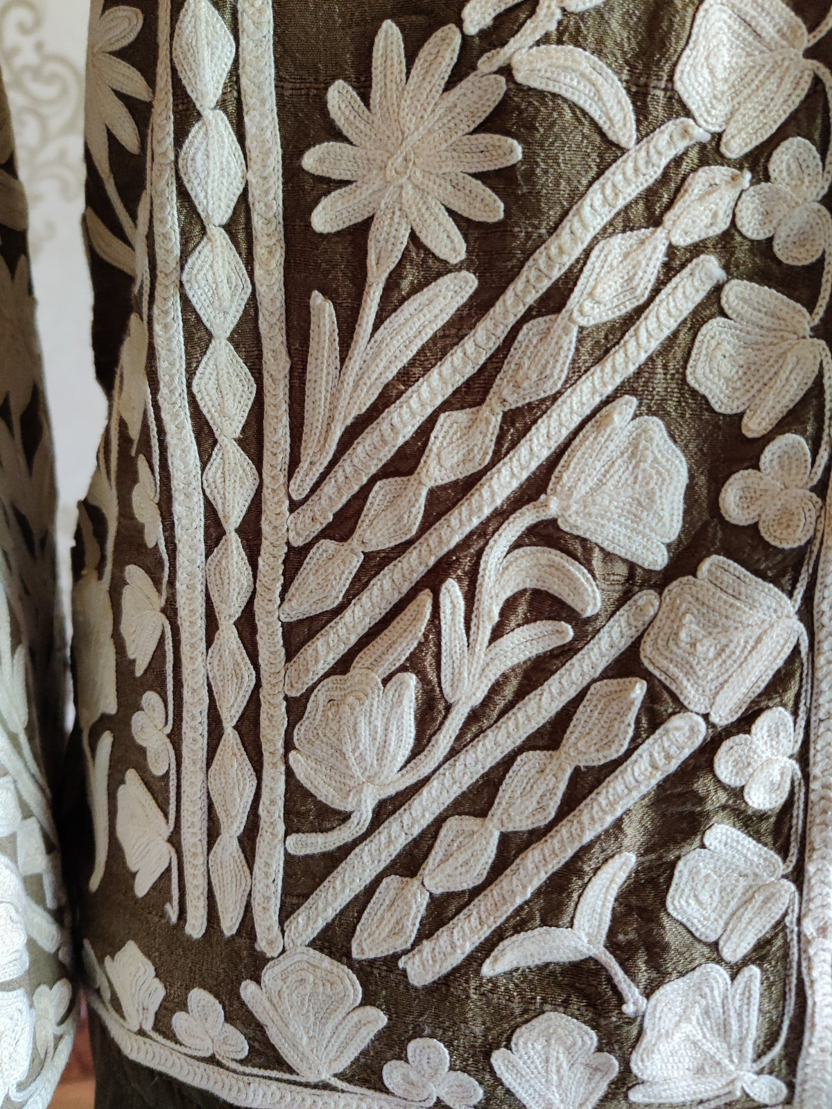 Close-up of white Aari flowers, leaves and diagonal decorative bands near the brown jacket’s lower edge.