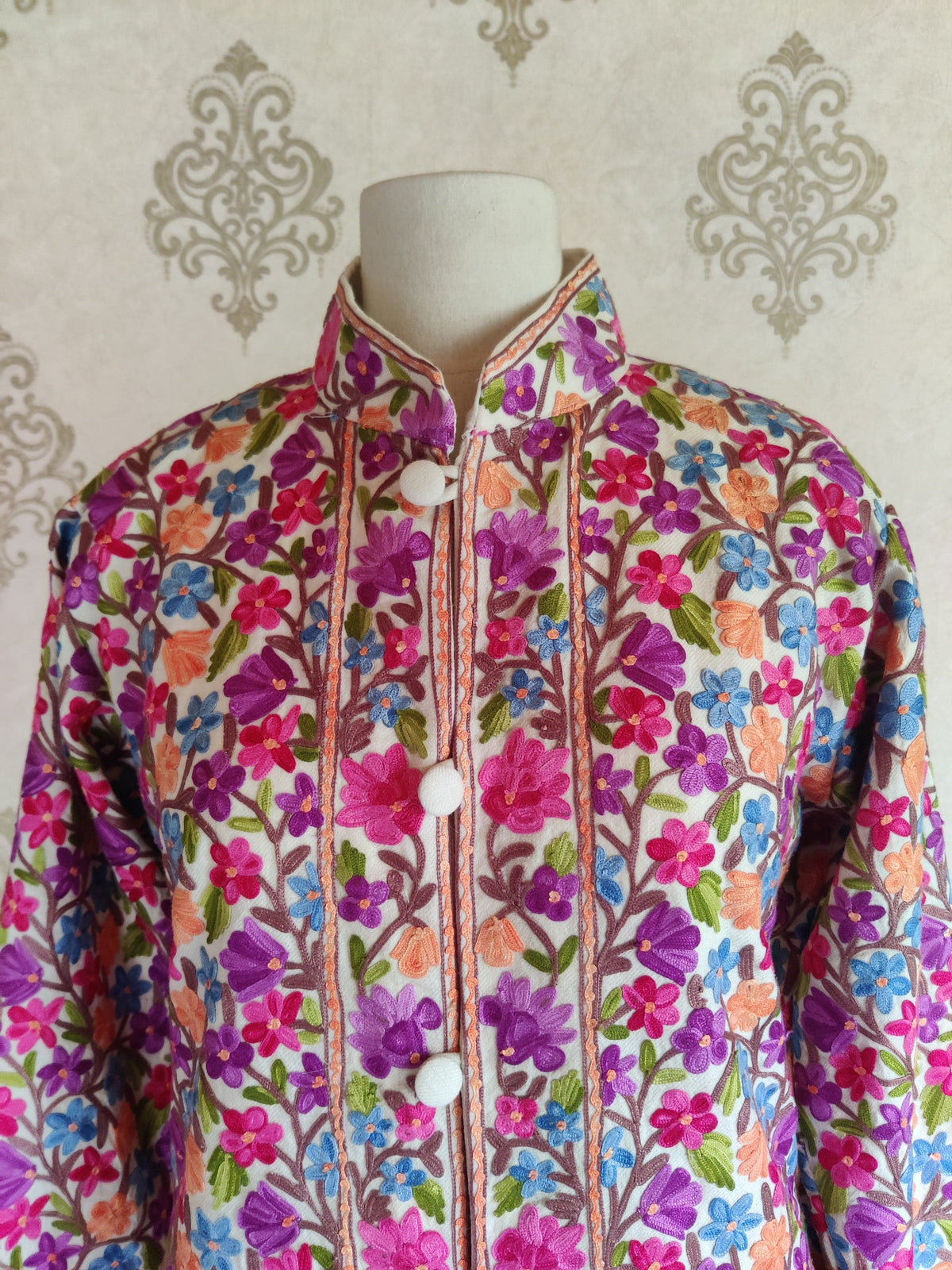 Upper front of a white jacket with pink and purple Aari flowers around the stand collar and buttoned opening