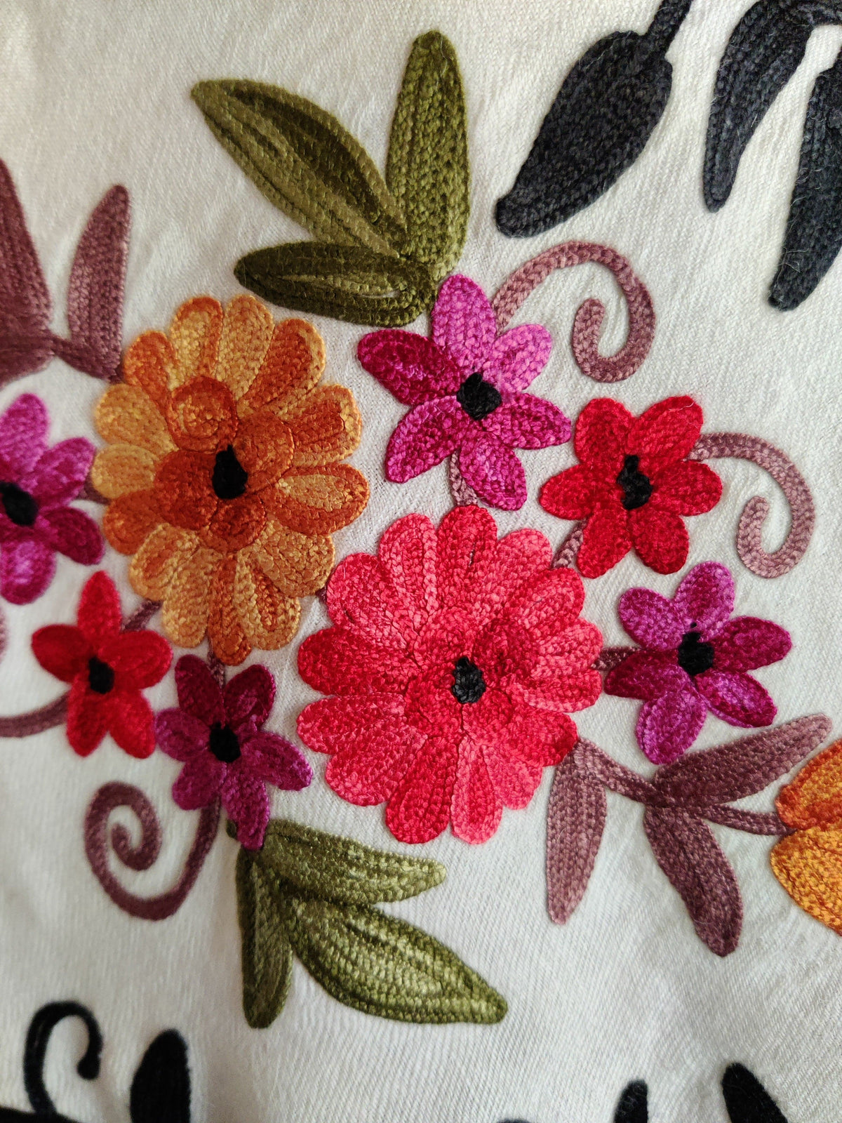 Close-up of red, pink and orange Aari flowers with green leaves, mauve curling stems and black leaves on a white merino wool stole.