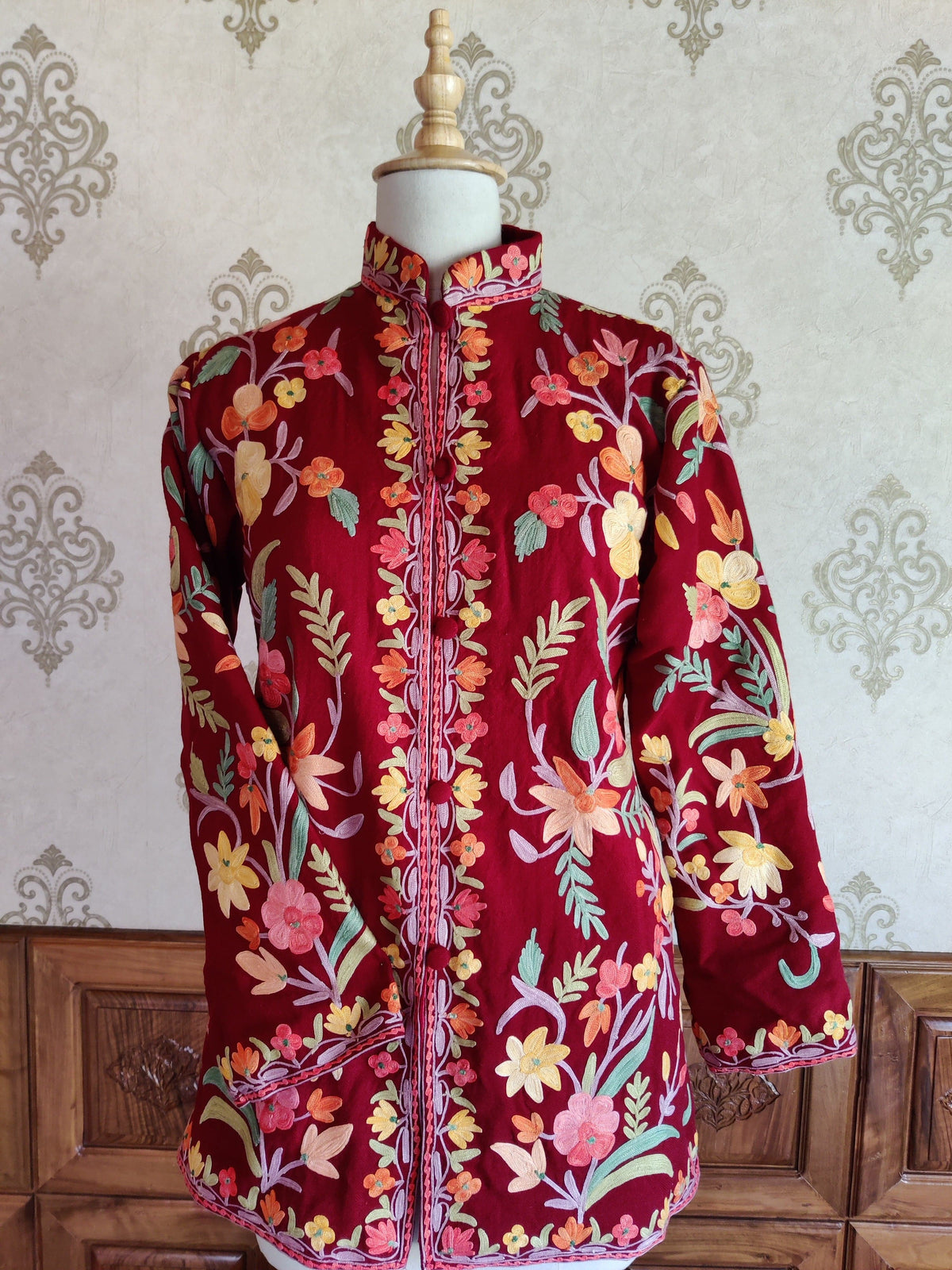 Front of a short maroon Aari jacket with pastel floral vines, a stand collar and one sleeve folded across the waist