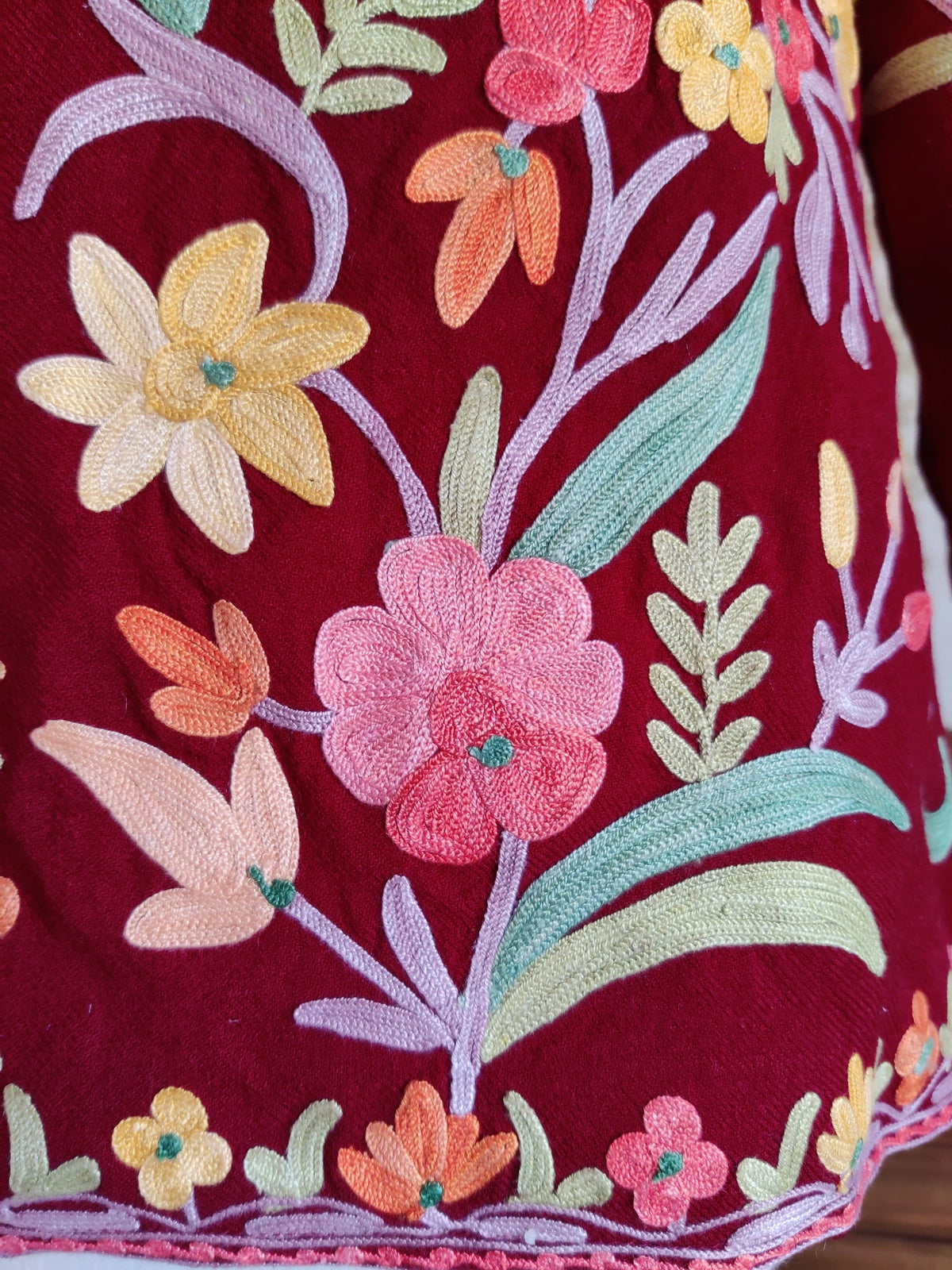 Detail of pink, yellow and peach Aari flowers on lilac stems with mint leaves above the floral hem of a maroon jacket