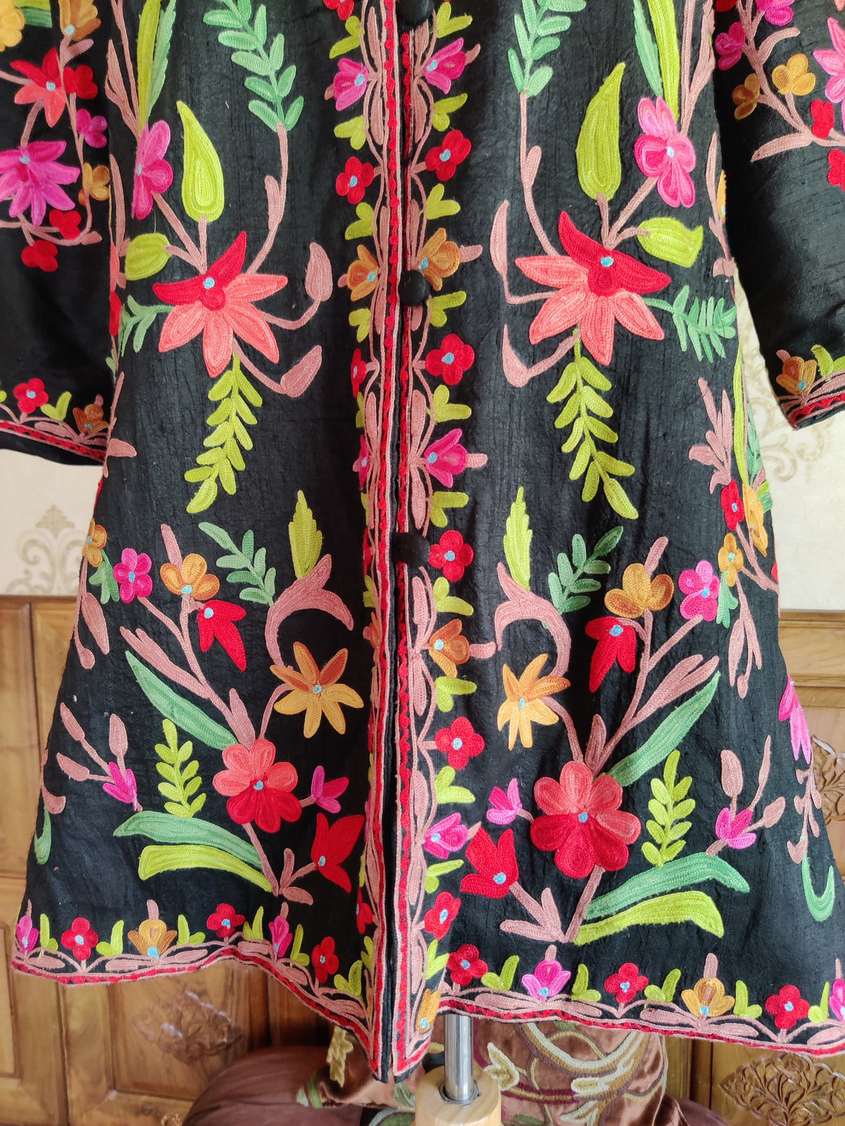 Lower front detail of a black jacket with bright red and pink Aari flowers and a narrow floral border beside the buttons