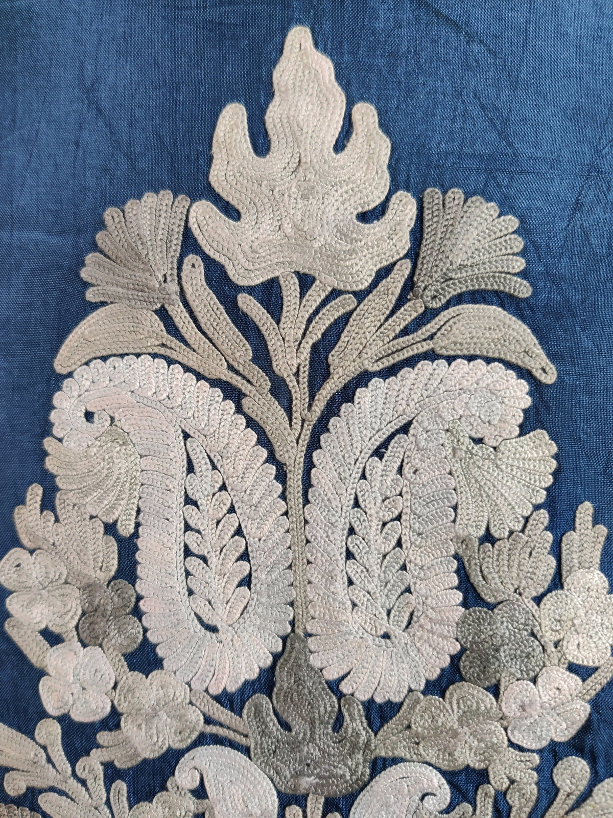 Close-up of white and silver-grey paisleys, flowers and leaves embroidered on blue fabric.