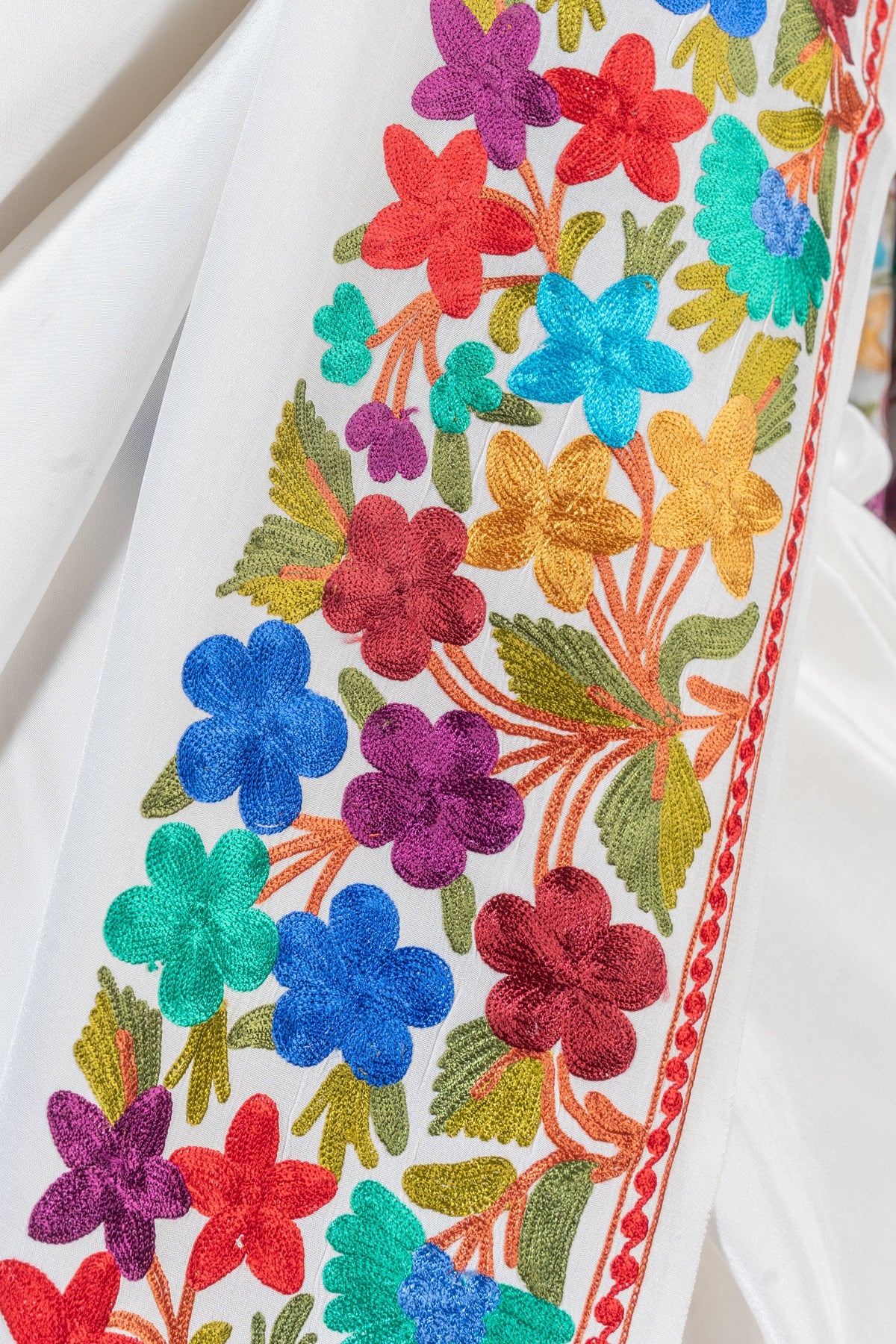 Close-up of the white saree border with bright multicoloured Aari flowers, green leaves and narrow red edging