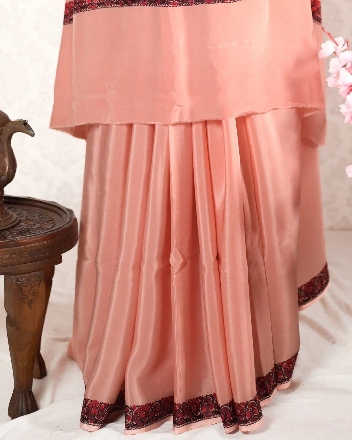 Lower pleats of a peach crepe saree with a narrow red-and-dark embroidered floral border at the hem.