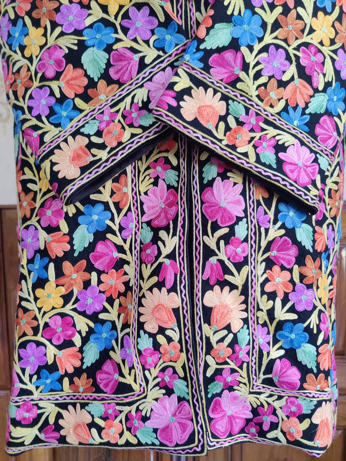 Detail of crossed cuffs and the lower front of the black Aari jacket, with colourful flowers framed by narrow decorative lines.