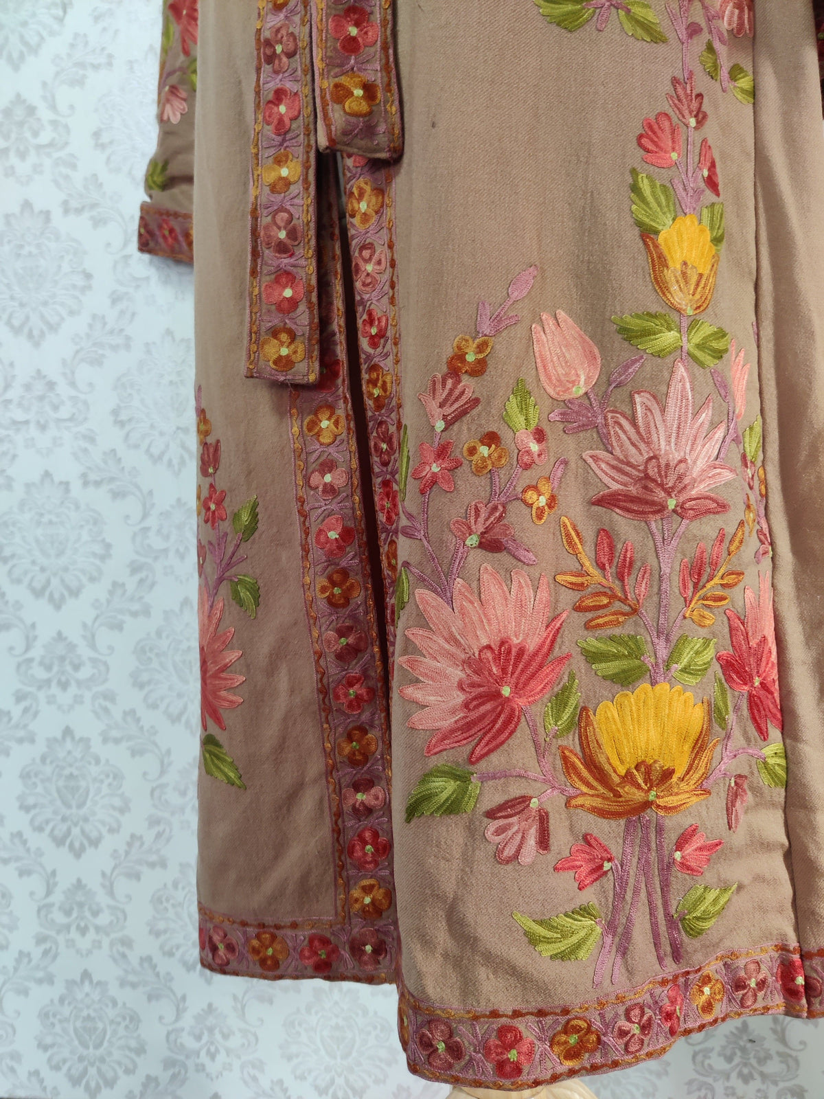 Lower front detail of a taupe-brown robe with large pink and yellow floral bouquets beside an embroidered border.