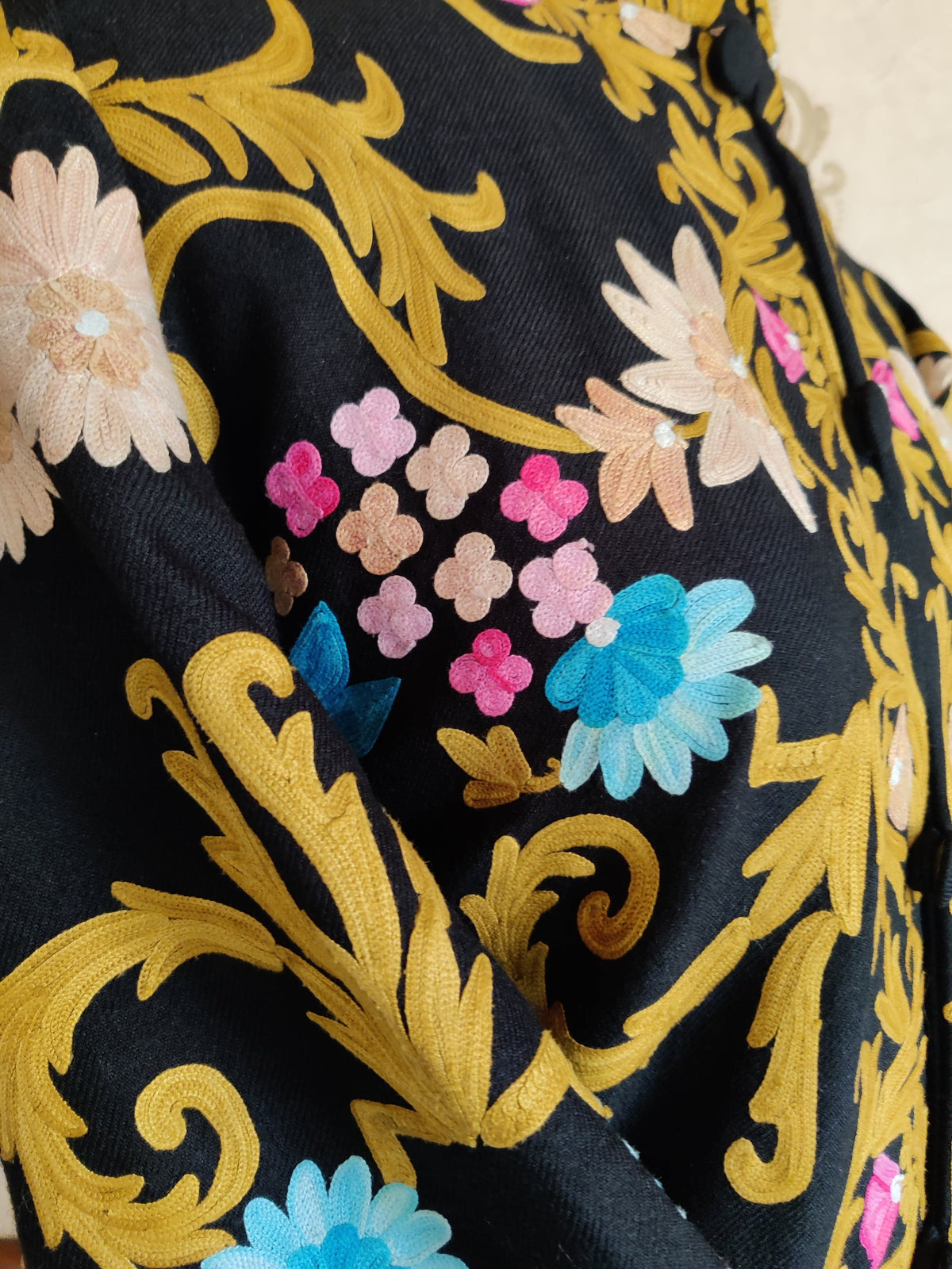 Shoulder detail of a black coat with golden Aari vines, blue flowers and small clustered pink and beige blossoms