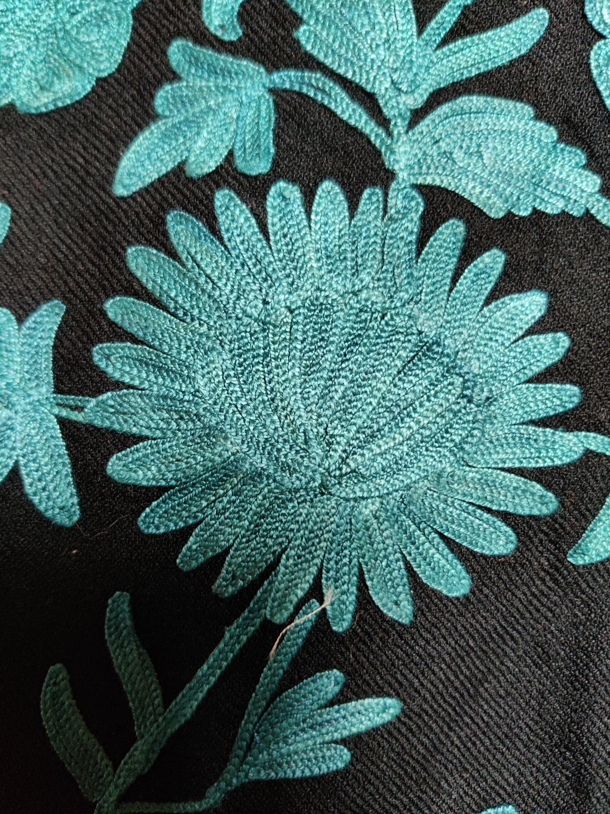 Macro detail of a turquoise Aari flower and leaves embroidered on the black jacket fabric.