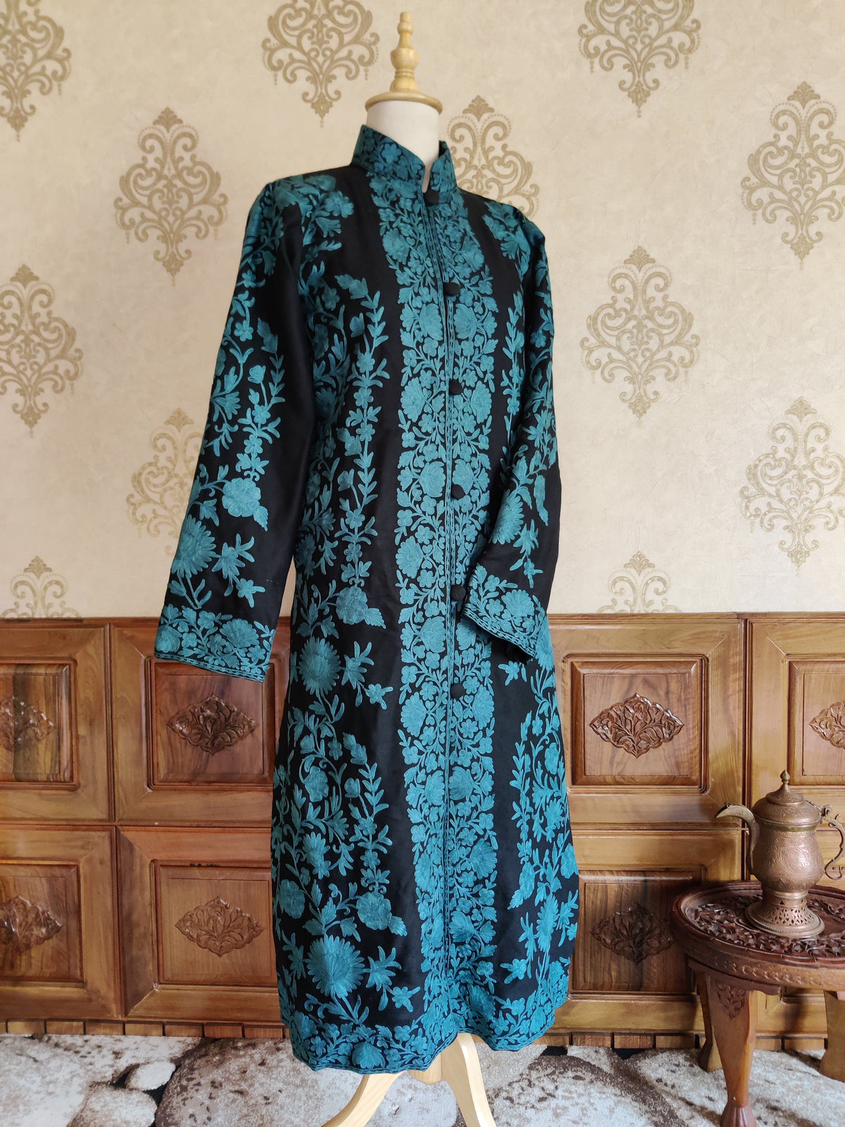 Three-quarter front of the long black jacket on a mannequin, showing turquoise Aari floral embroidery and a standing collar.