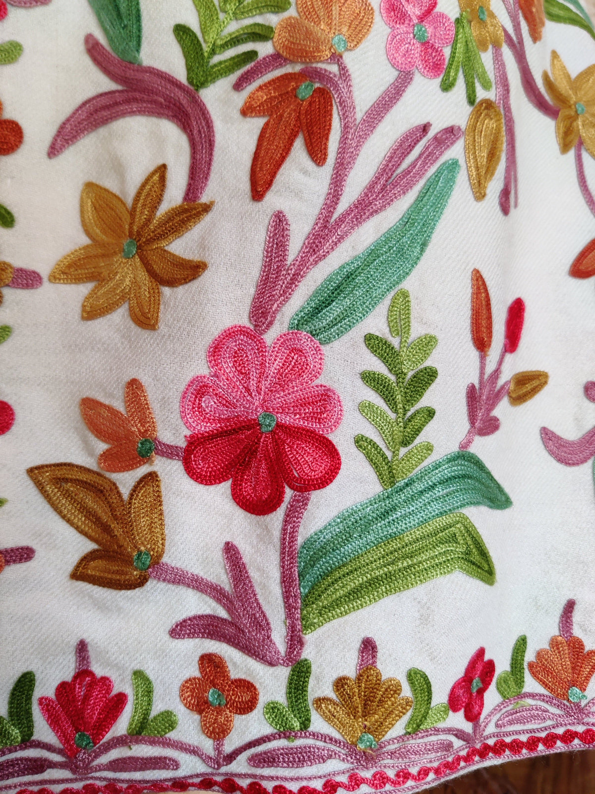 Detail of pink and red Aari flowers, teal and green leaves and a narrow floral hem border on a white jacket
