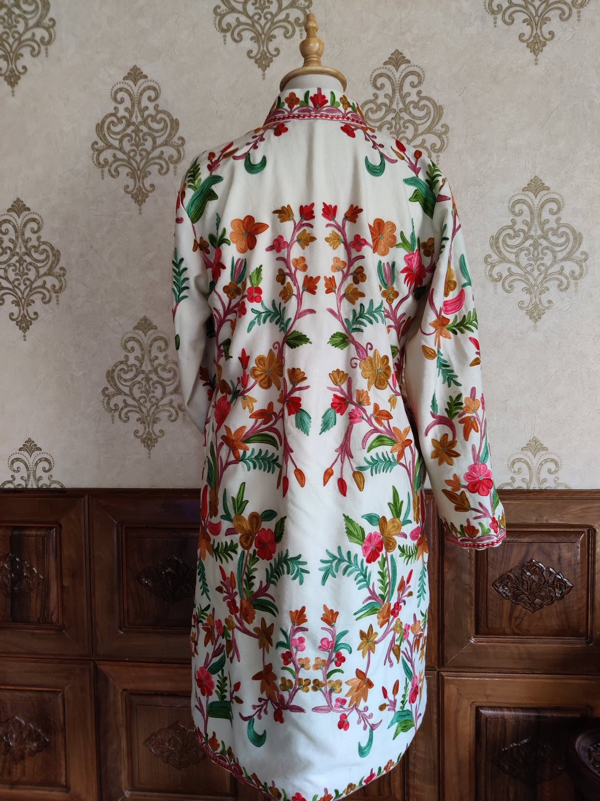 Back of a long white Aari jacket on a mannequin, with symmetrical multicoloured floral vines and embroidered cuffs