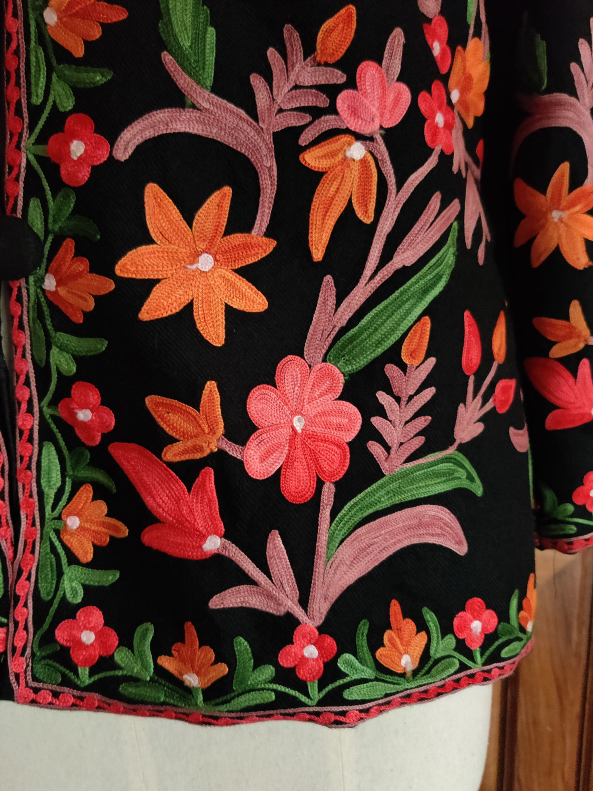 Lower front detail of a black jacket with bright orange and pink Aari flowers above a narrow floral hem border