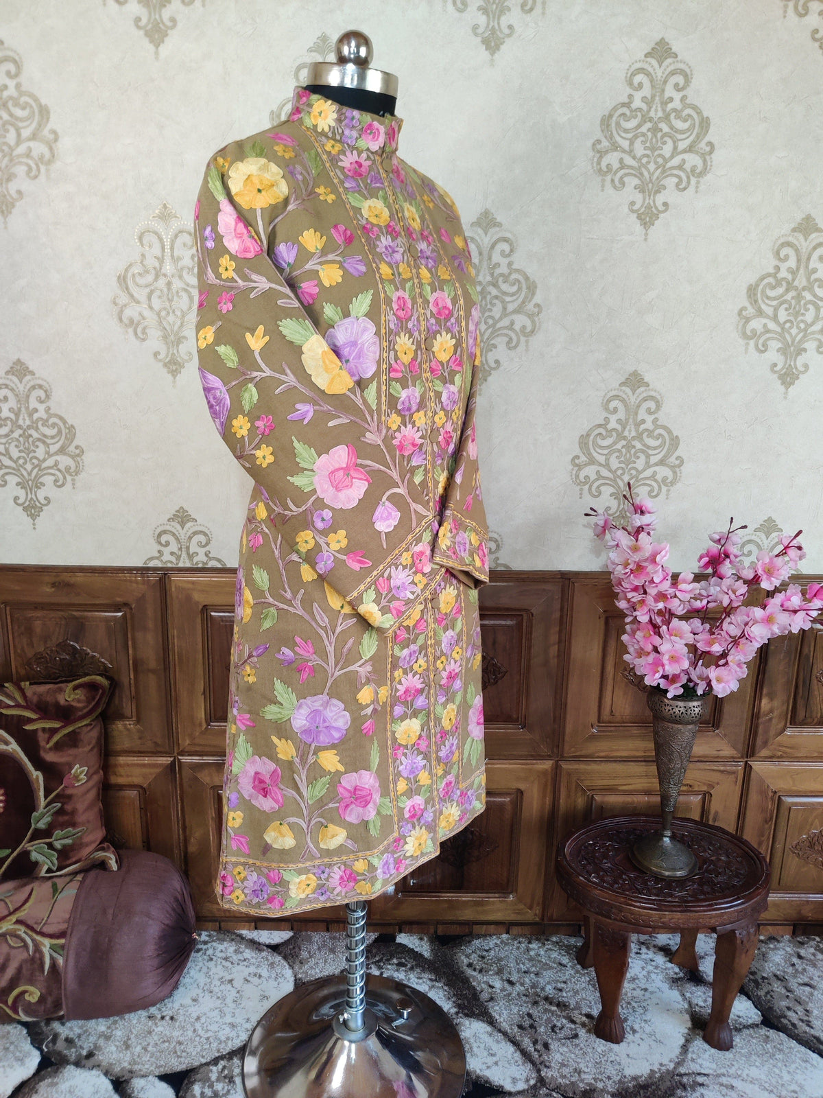 Three-quarter view of taupe long coat with crossed sleeves, showing colourful Aari floral vines and an embroidered hem