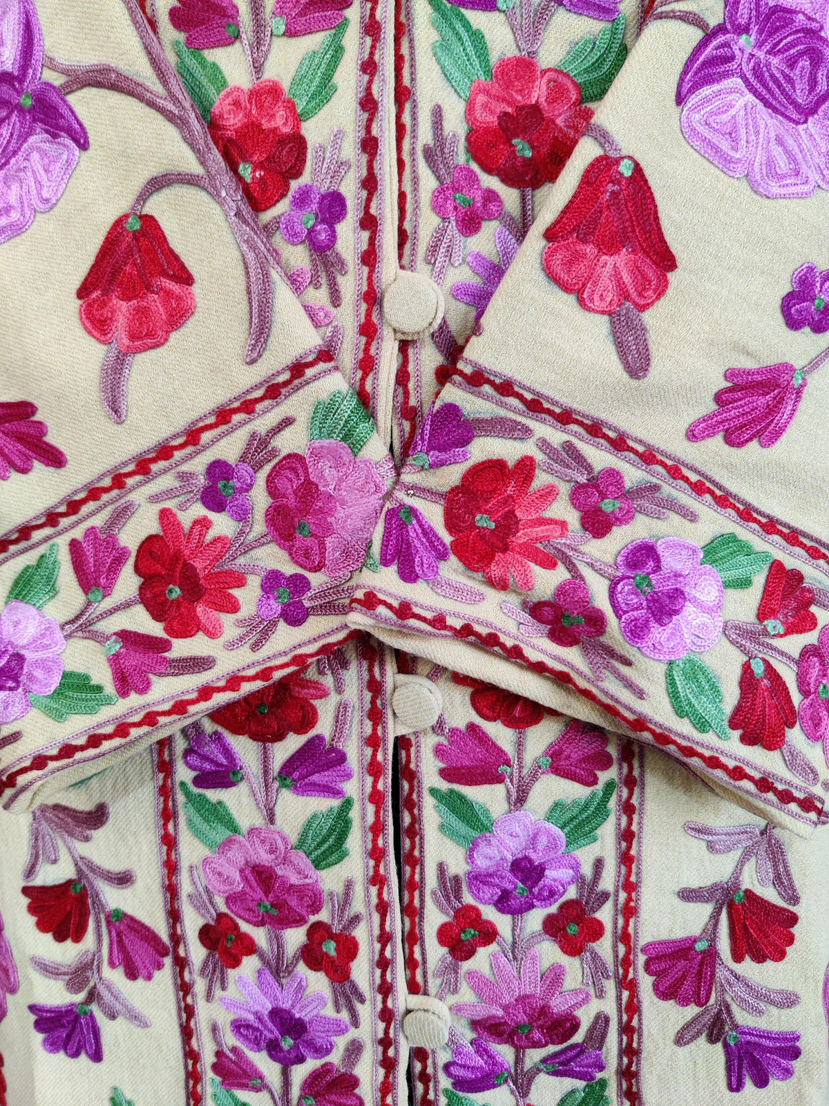 Close-up of crossed cuffs and covered buttons on a cream jacket, embroidered with bright pink, red and purple Aari flowers