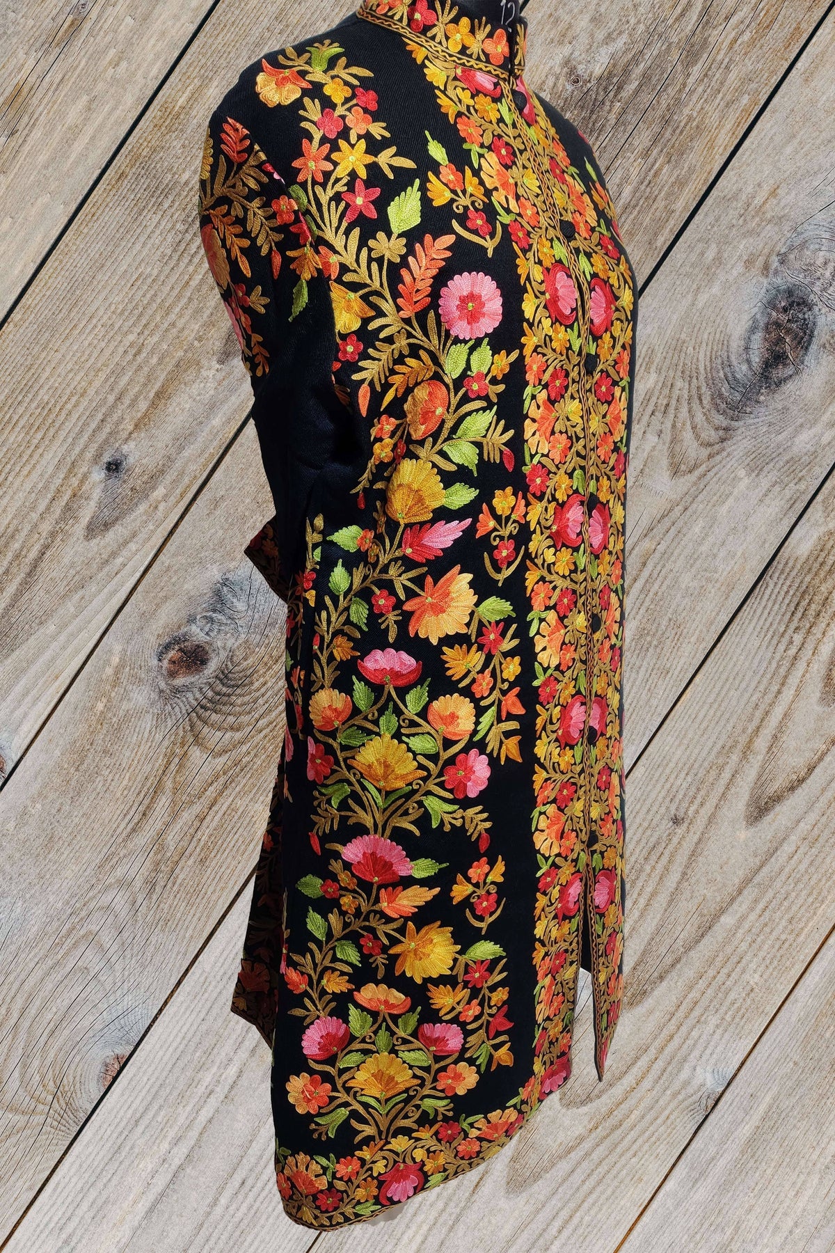 Side-front view of a black long jacket with pink and orange Aari floral vines down the sleeve and side, beside a dense floral placket