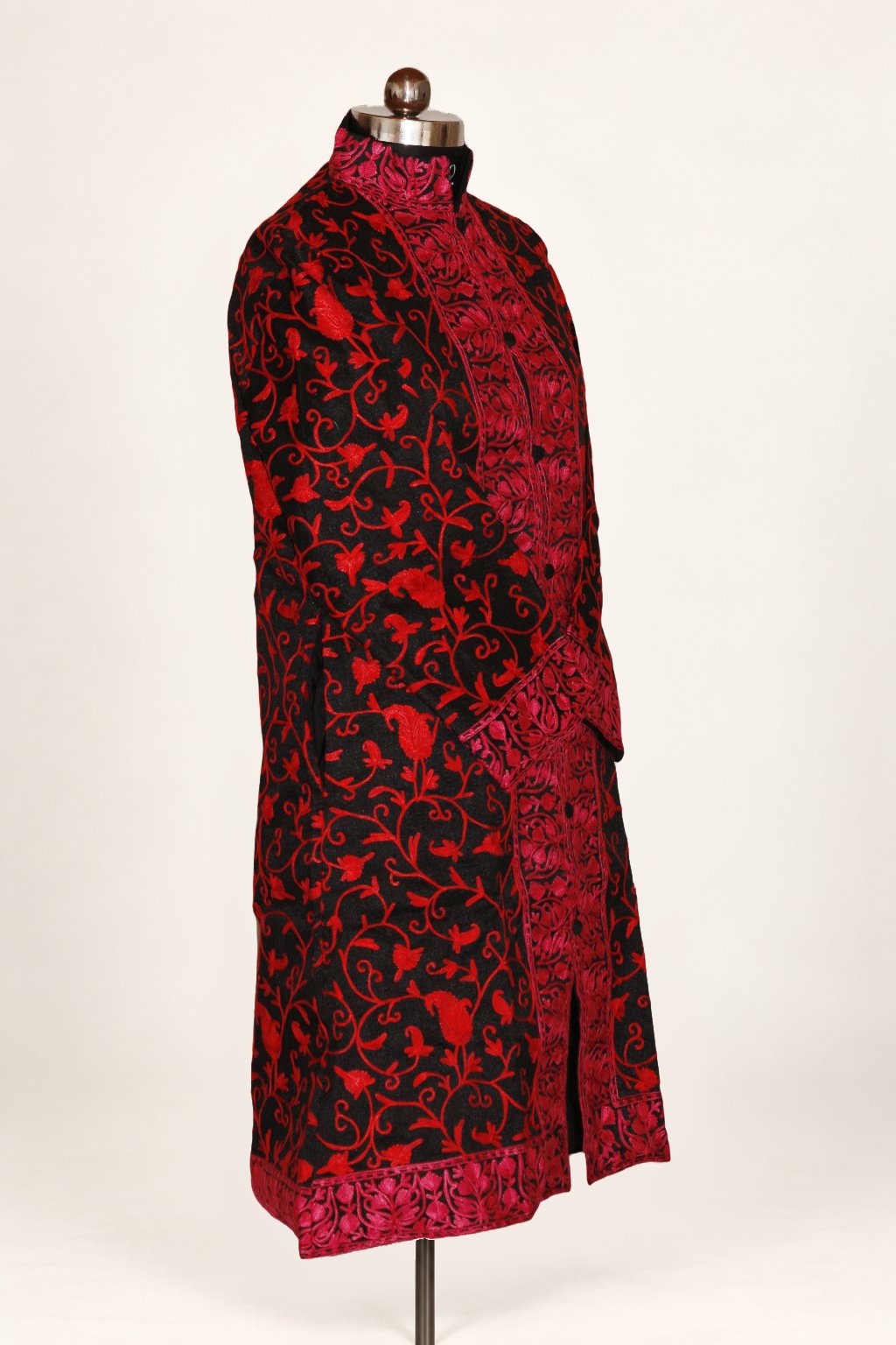 Three-quarter view of black long coat on a mannequin, with red Aari floral vines and dense collar, front and hem borders