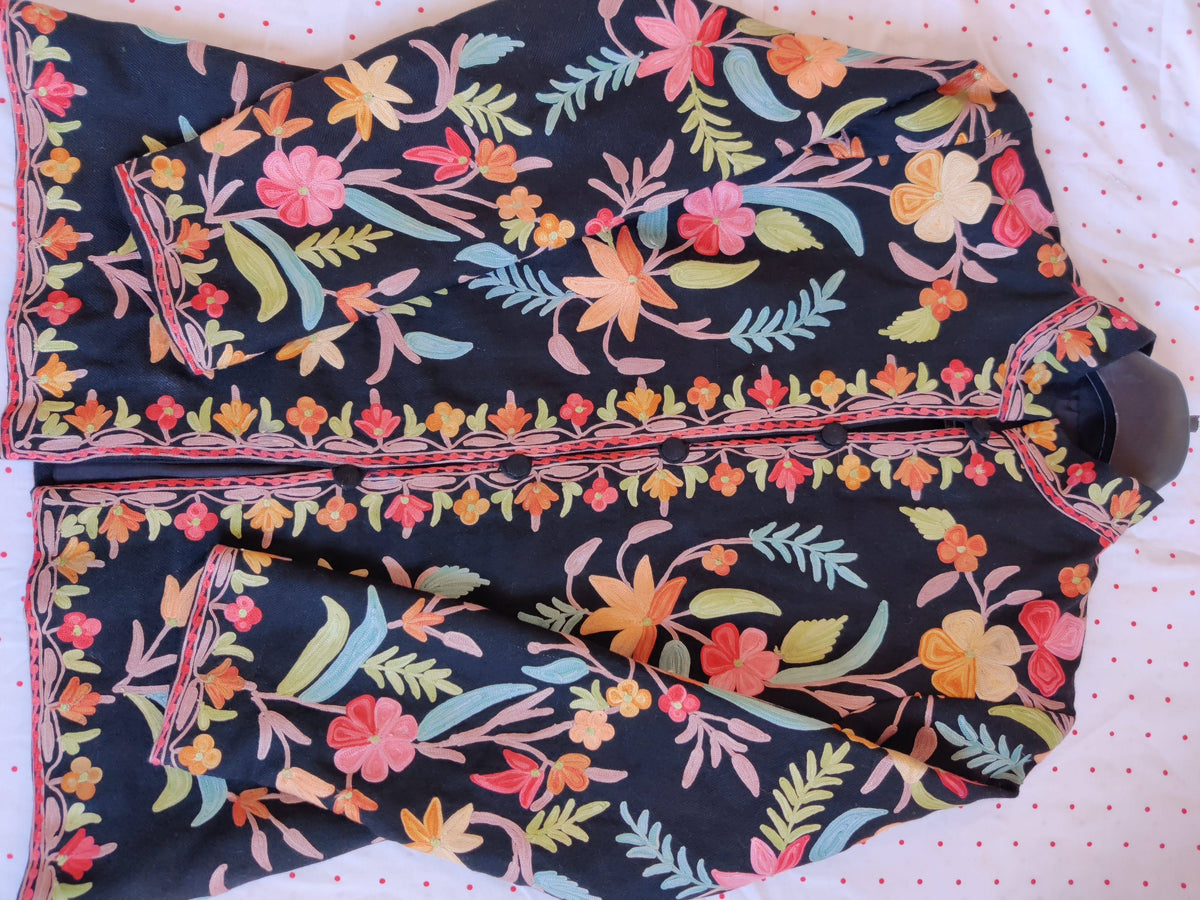 Black short jacket laid sideways with sleeves folded inward, showing colourful Aari embroidery and floral front borders