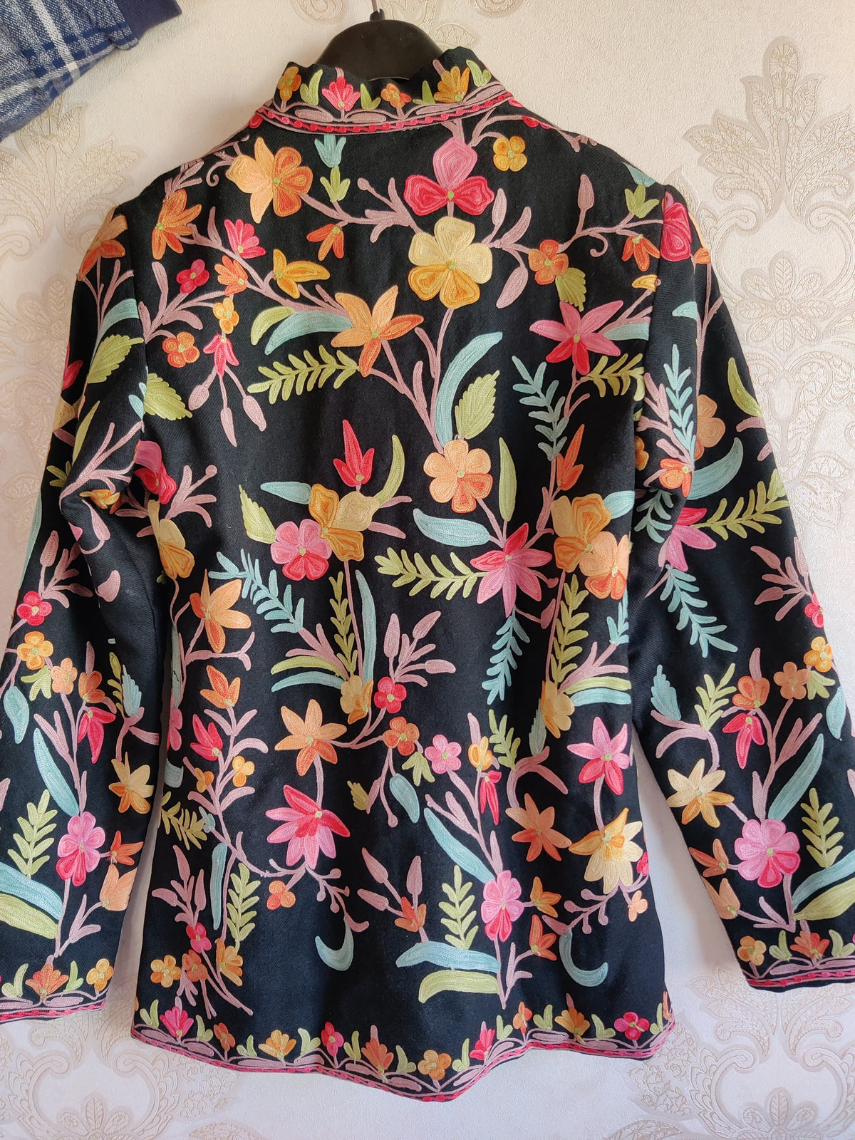 Back of black short jacket on a hanger, with pink, orange and yellow Aari flowers and pale blue and green leaves