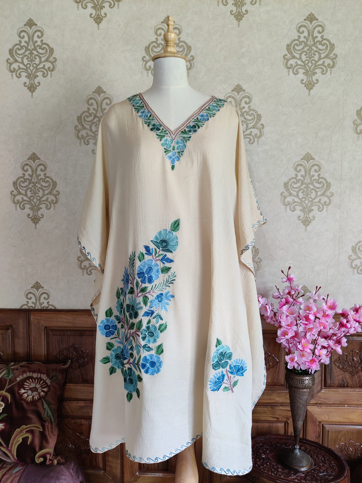 Front of a cream kaftan on a mannequin, with a blue-green Aari V-neck border, a large floral spray and a smaller motif near the hem.