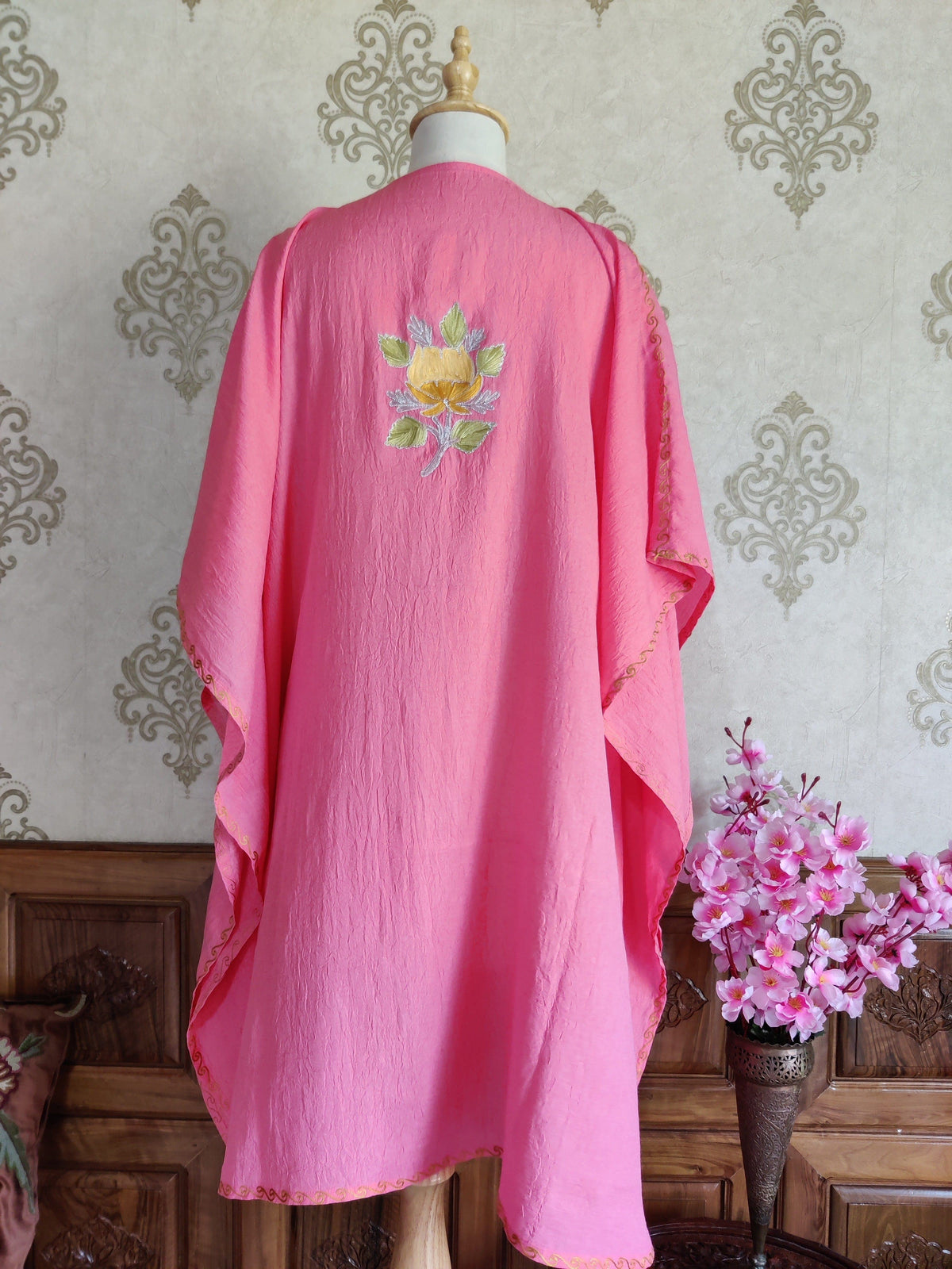Back of a pink kaftan on a mannequin, with one yellow floral motif and green leaves between the shoulders and wide draped sleeves.