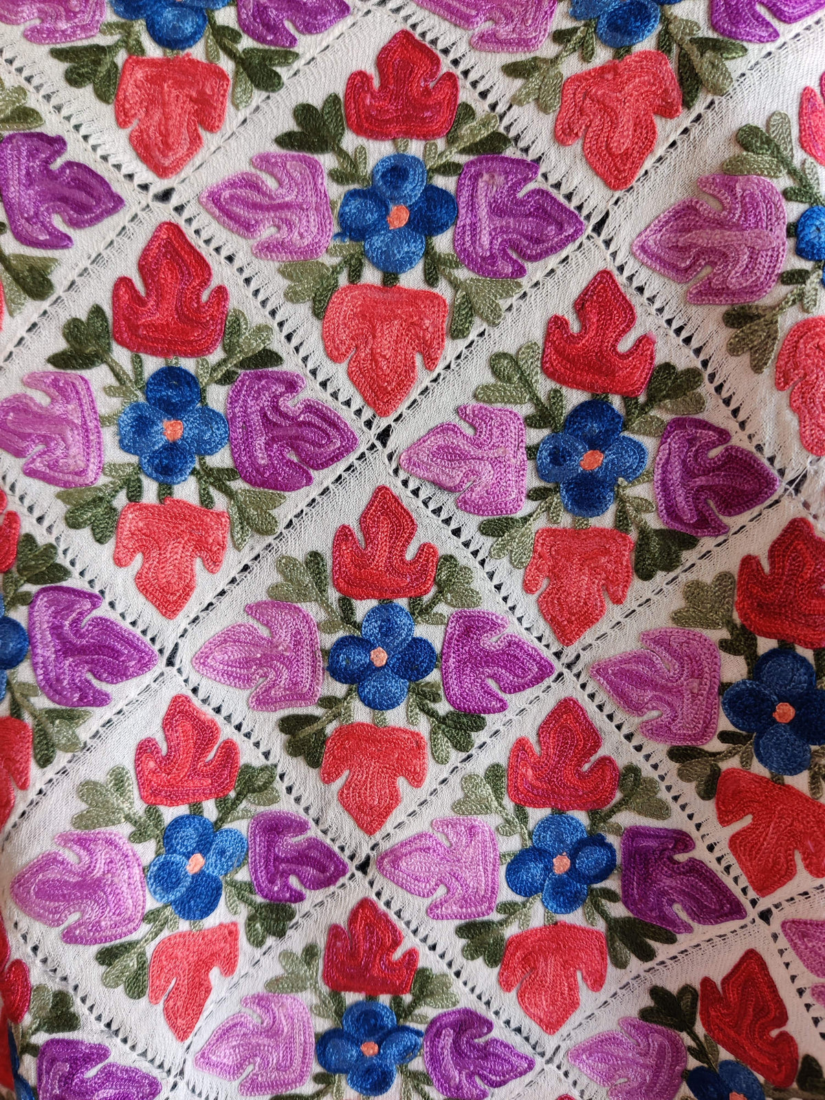 Close-up of pink and purple Aari leaf shapes around blue flowers, separated by white cutwork lines on a Merino wool stole