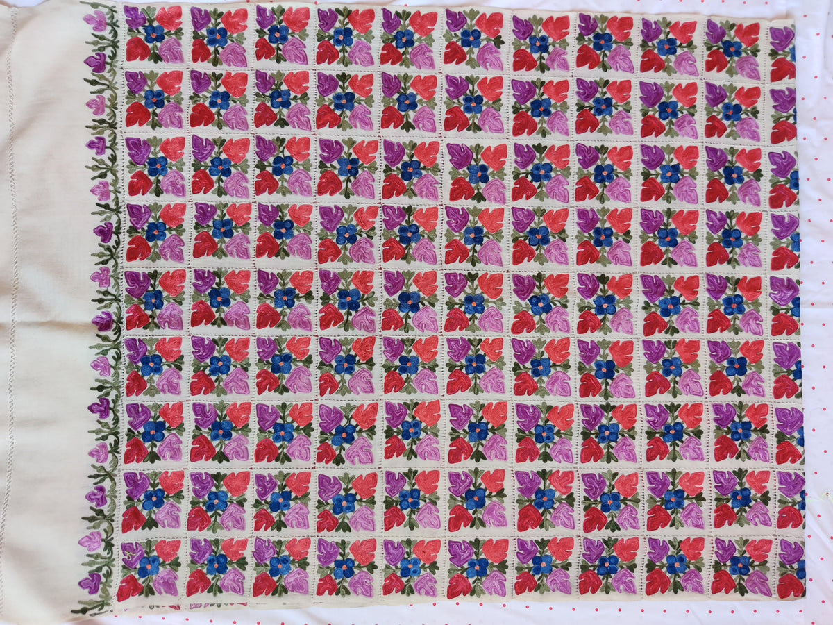 White Merino wool stole laid flat, with a regular grid of pink, purple and blue Aari floral motifs and a narrow leafy border