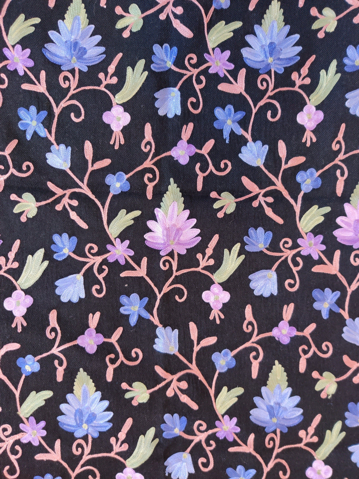 Close-up of blue and lavender Aari flowers with pink curling stems and pale green leaves across a black merino wool stole.