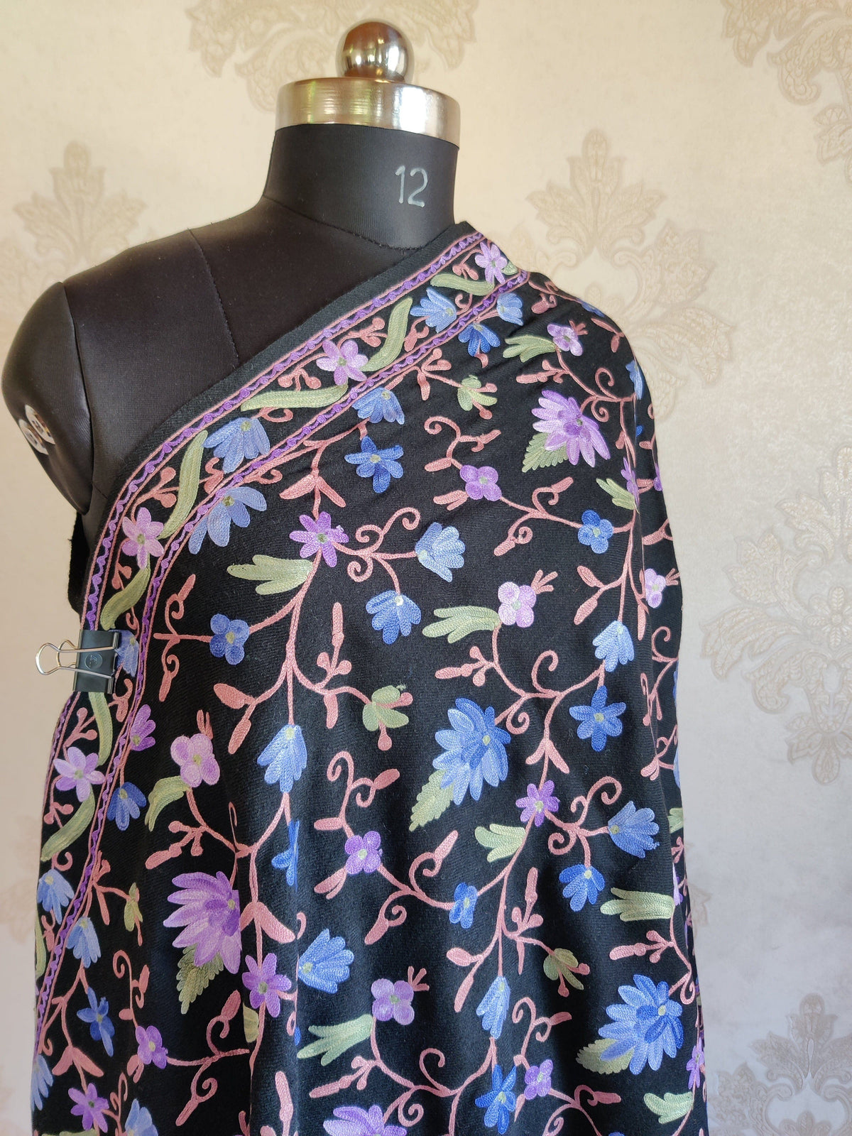 Upper view of a black merino wool stole across a mannequin's chest, with blue and lavender Aari flowers and a purple-edged floral border.