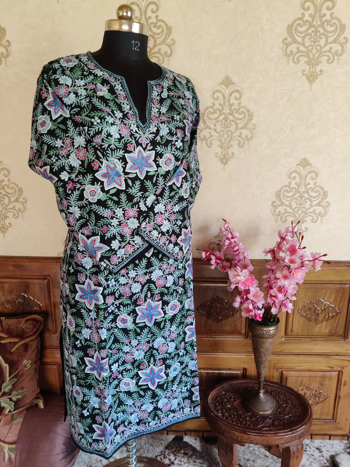 Full-length black pheran on a mannequin with sleeves crossed, showing dense pastel Aari floral embroidery and a split neckline