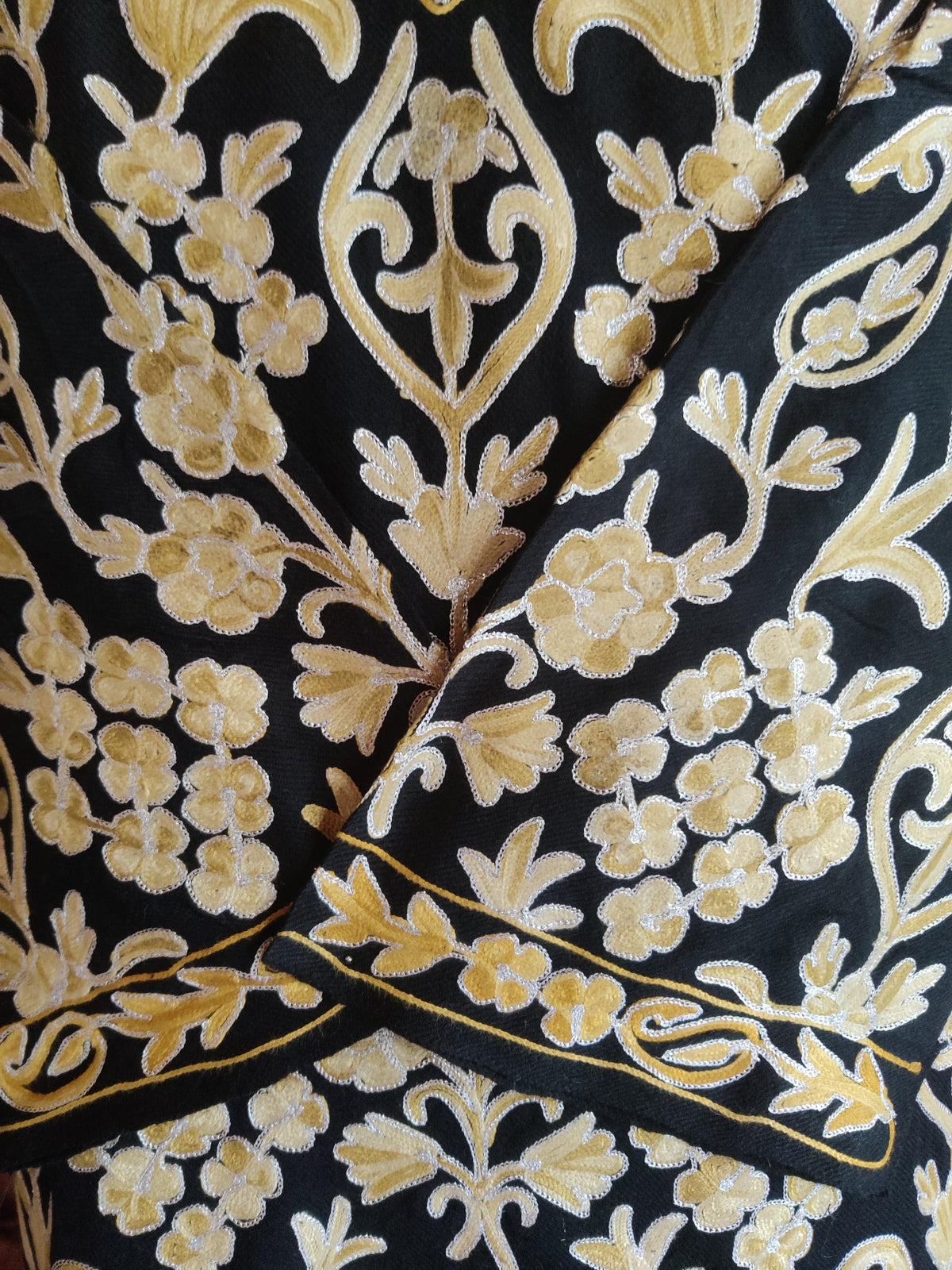 Close-up of pale-gold Aari flowers and curling leaves on the black pheran, with an embroidered cuff across the front.