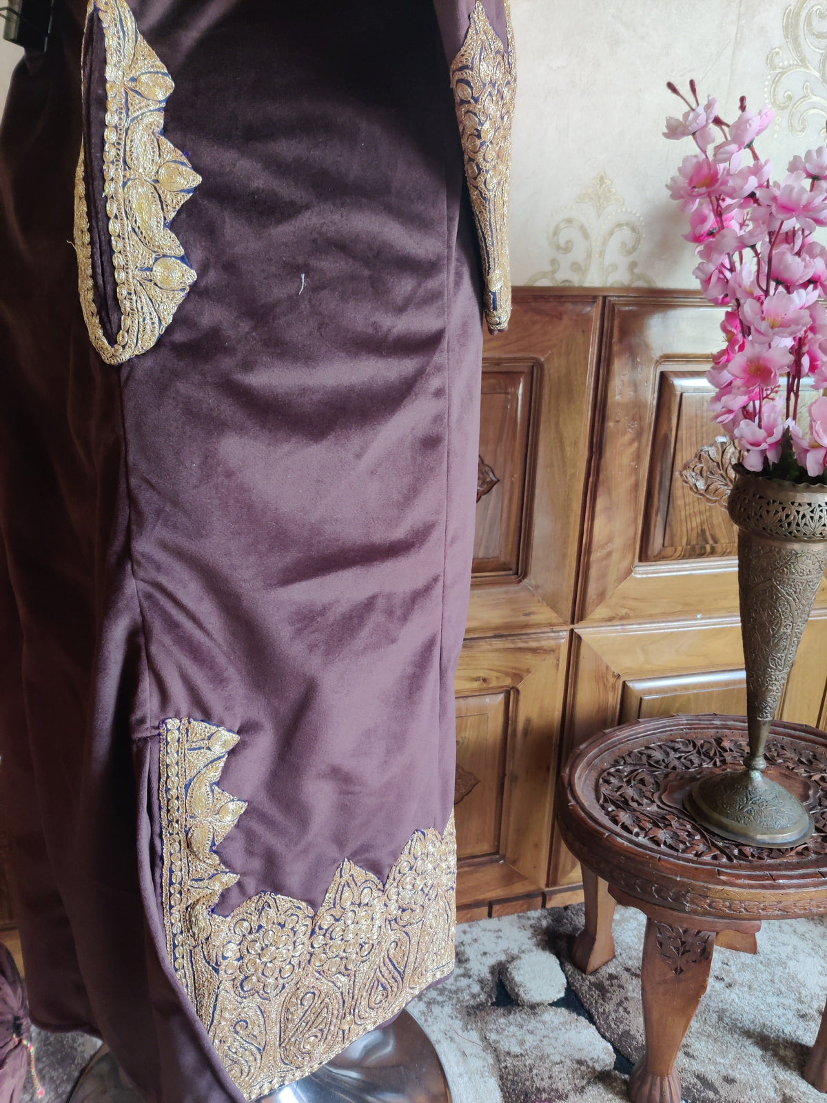 Side detail of a plum-burgundy velvet pheran, showing gold-toned Tilla trim around the pocket and side slit above a paisley hem border.