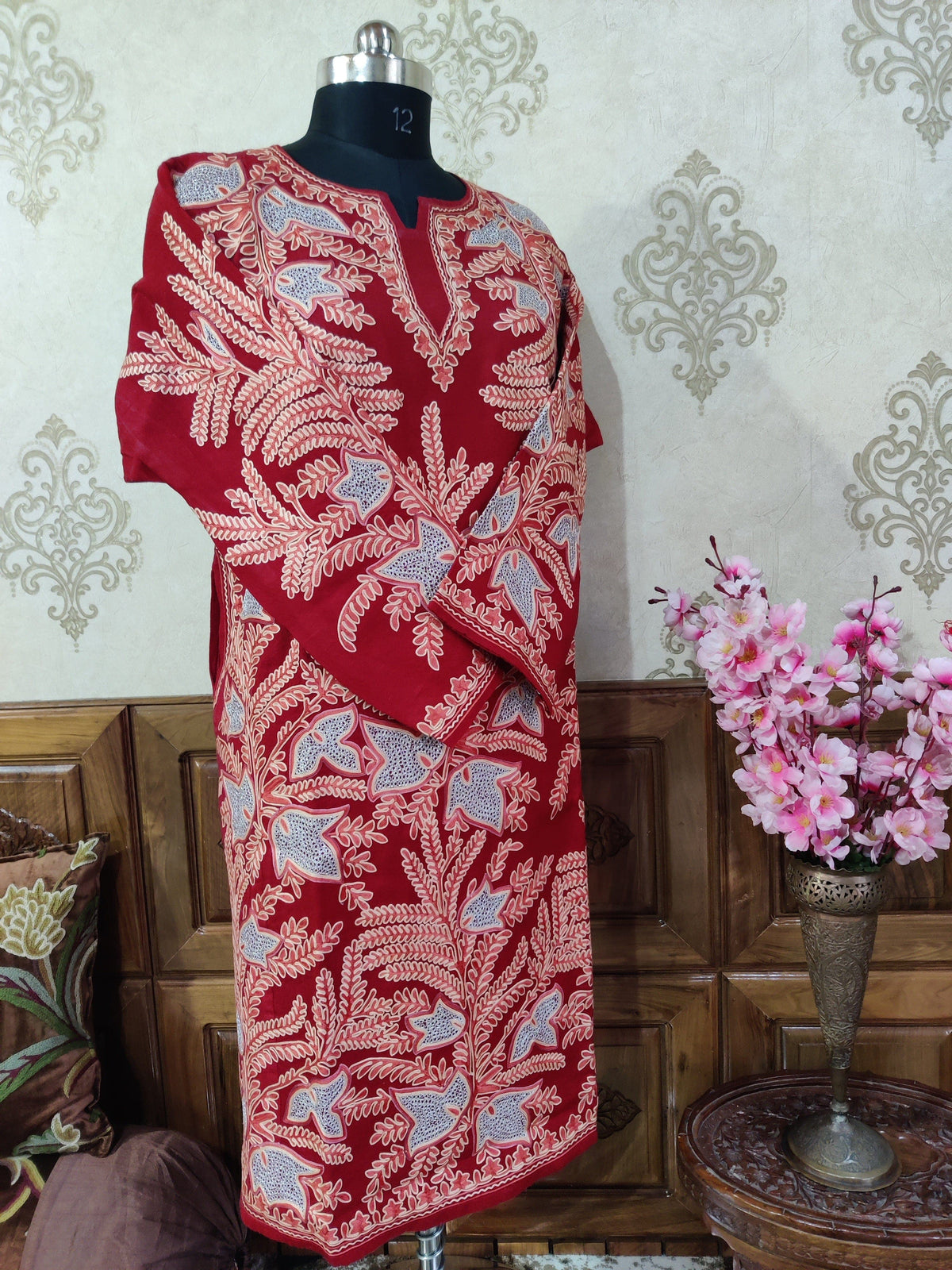 Angled full view of a red pheran covered in cream Aari leafy stems and pale blue Zari floral shapes, with sleeves crossed