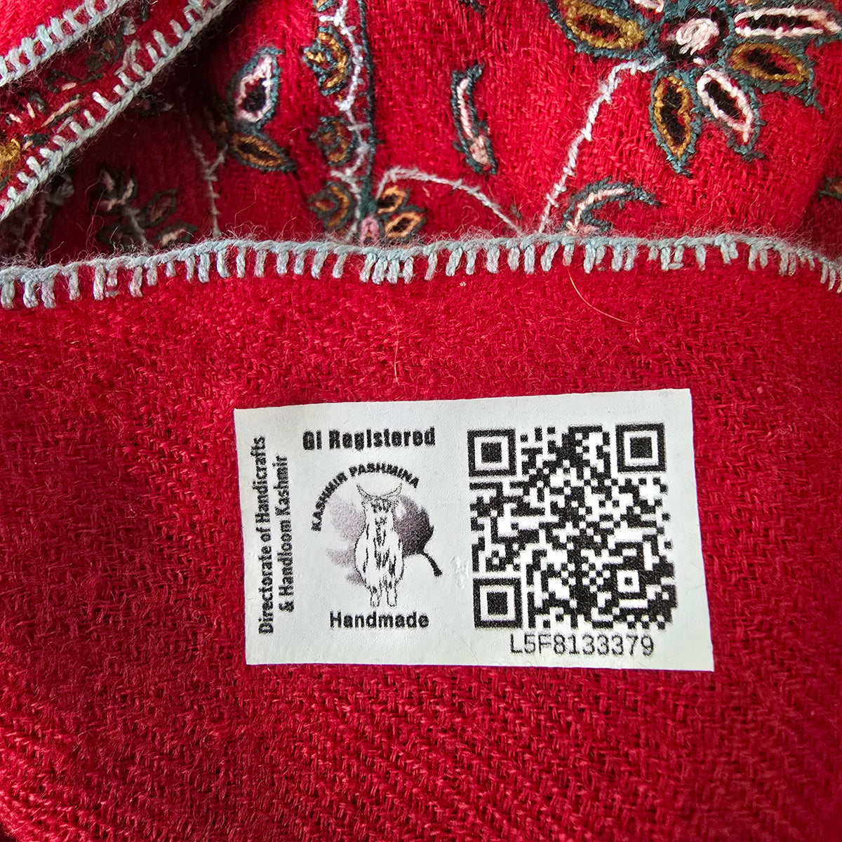 Close-up of a label marked GI Registered and Handmade, with a QR code, on scarlet embroidered shawl fabric