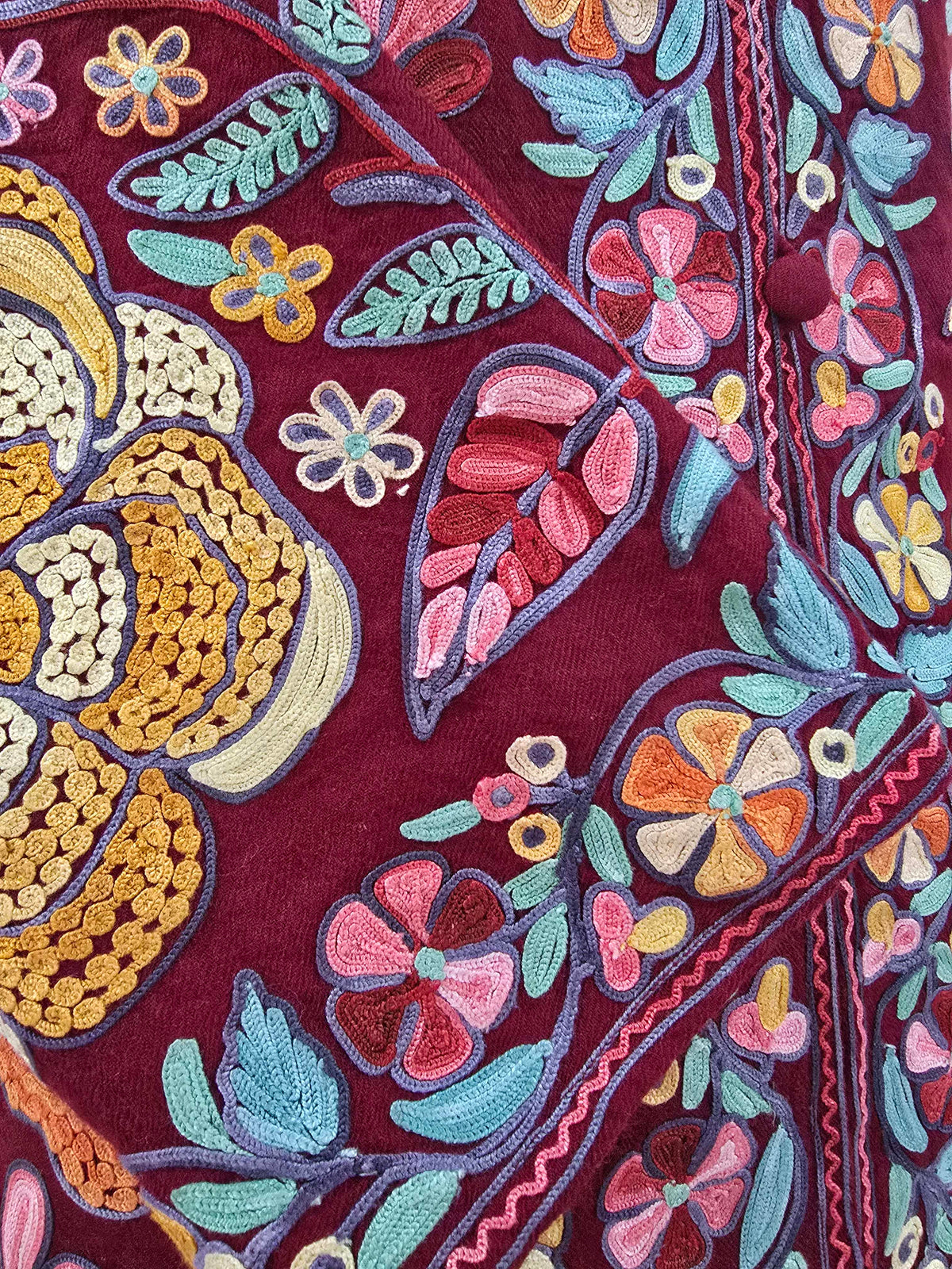 Close-up of pink, gold and teal floral Aari embroidery and a fabric-covered button on the burgundy jacket