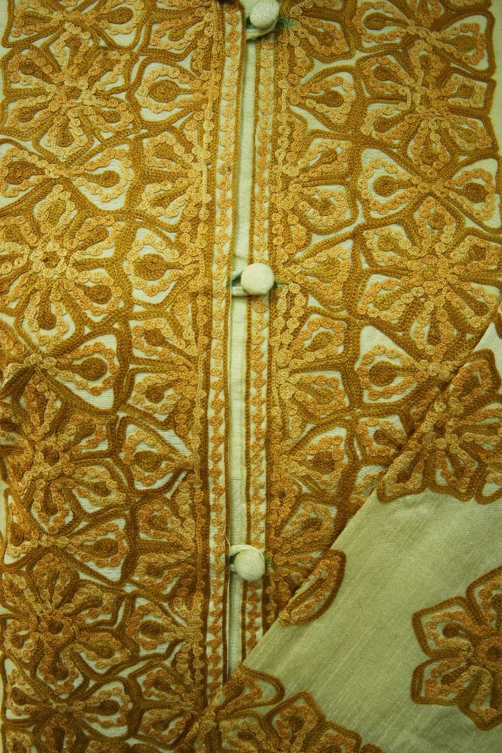 Detail of the ivory jacket’s covered buttons between dense gold Aari floral borders with small leaf-shaped elements.