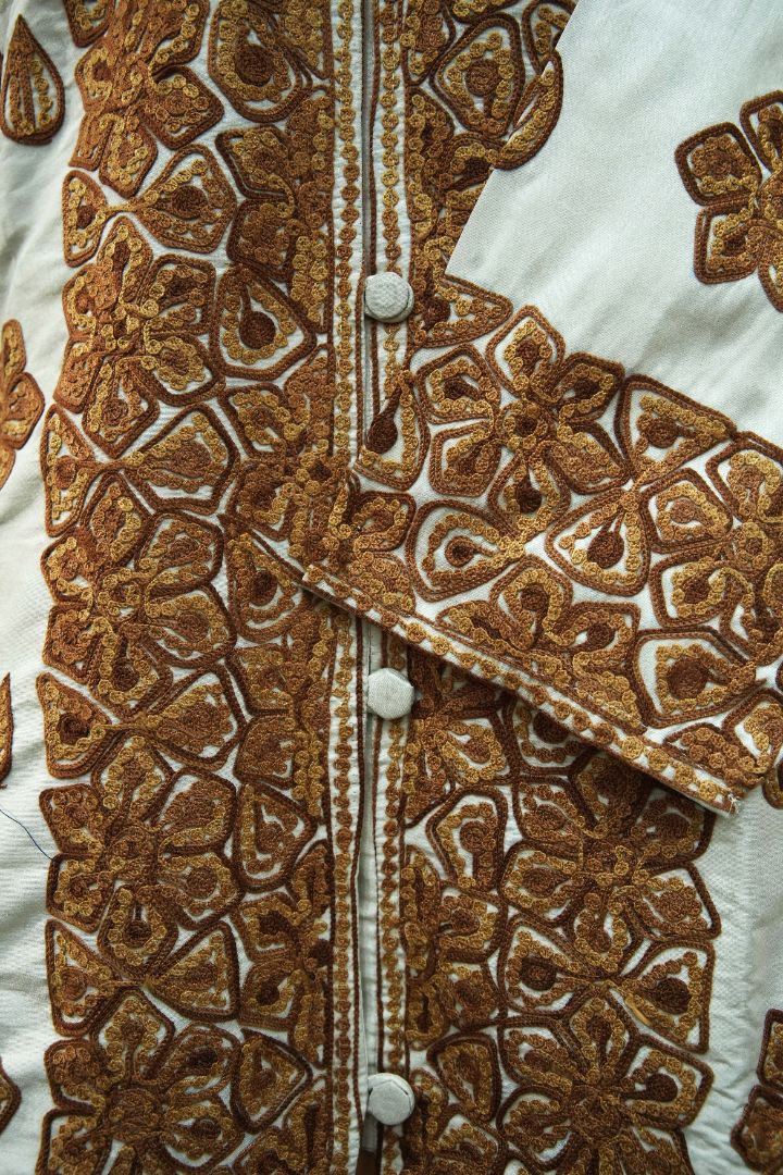 Close-up of the ivory jacket’s embroidered cuff and covered buttons beside broad copper-brown floral front borders.