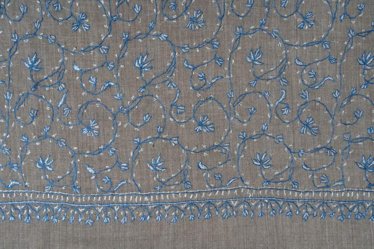 Grey shawl laid flat, showing blue and pale floral vines above narrow embroidered lines and small pendant leaf motifs
