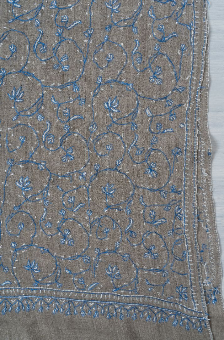 Corner of a grey shawl laid flat with blue Sozni vines, small flowers and a fine border along two edges