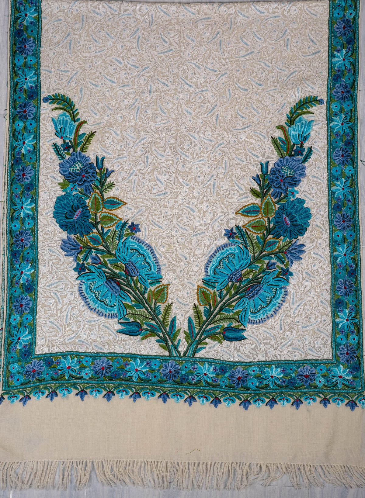 A square cream-colored textile, likely a shawl, scarf, or small rug, featuring a detailed floral pattern in shades of blue, teal, and green. The textile has a fringed edge on the right and is laid flat on a light gray tiled floor.