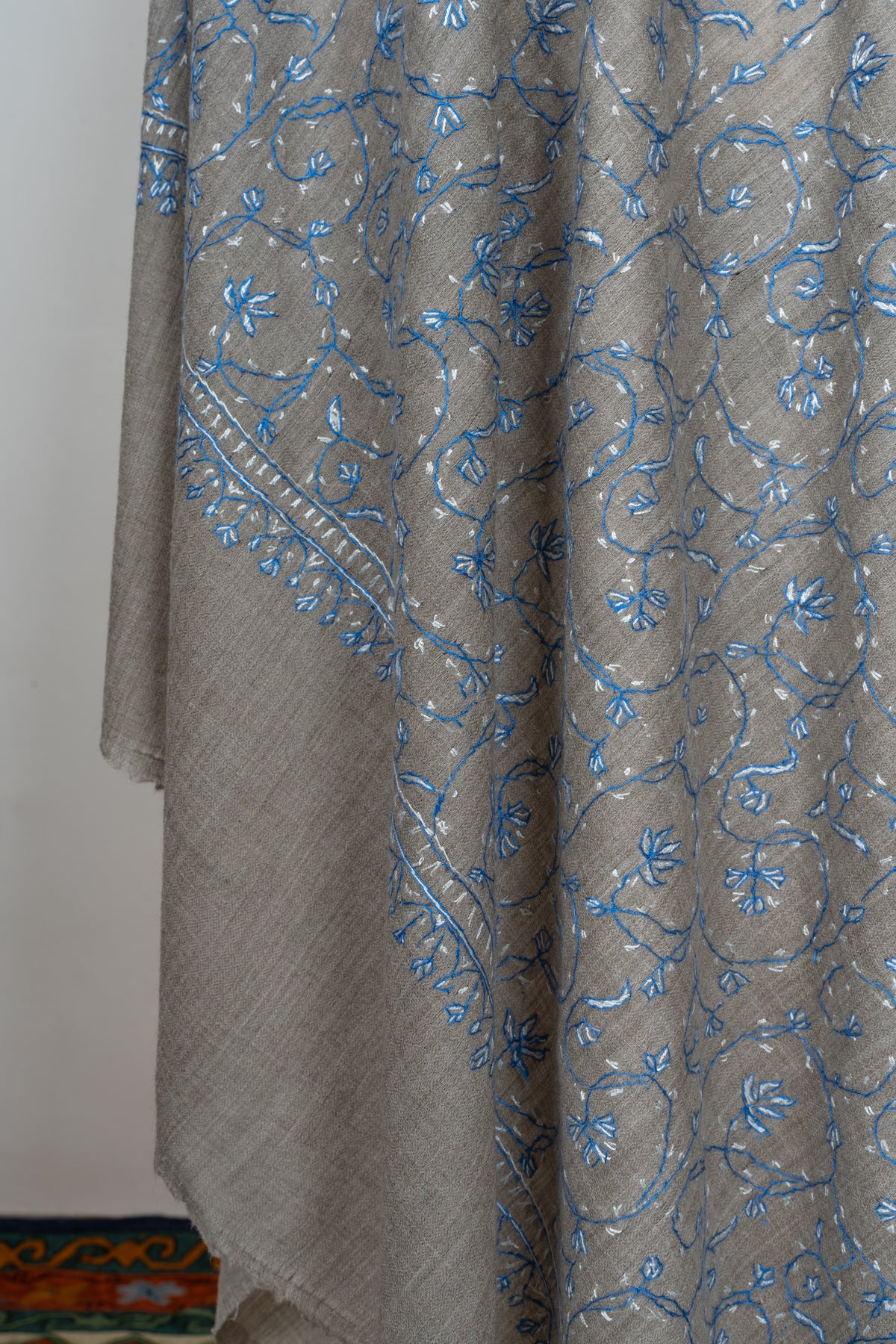 Hanging corner of a grey shawl with blue floral Sozni vines and fine embroidered edging above a plain end