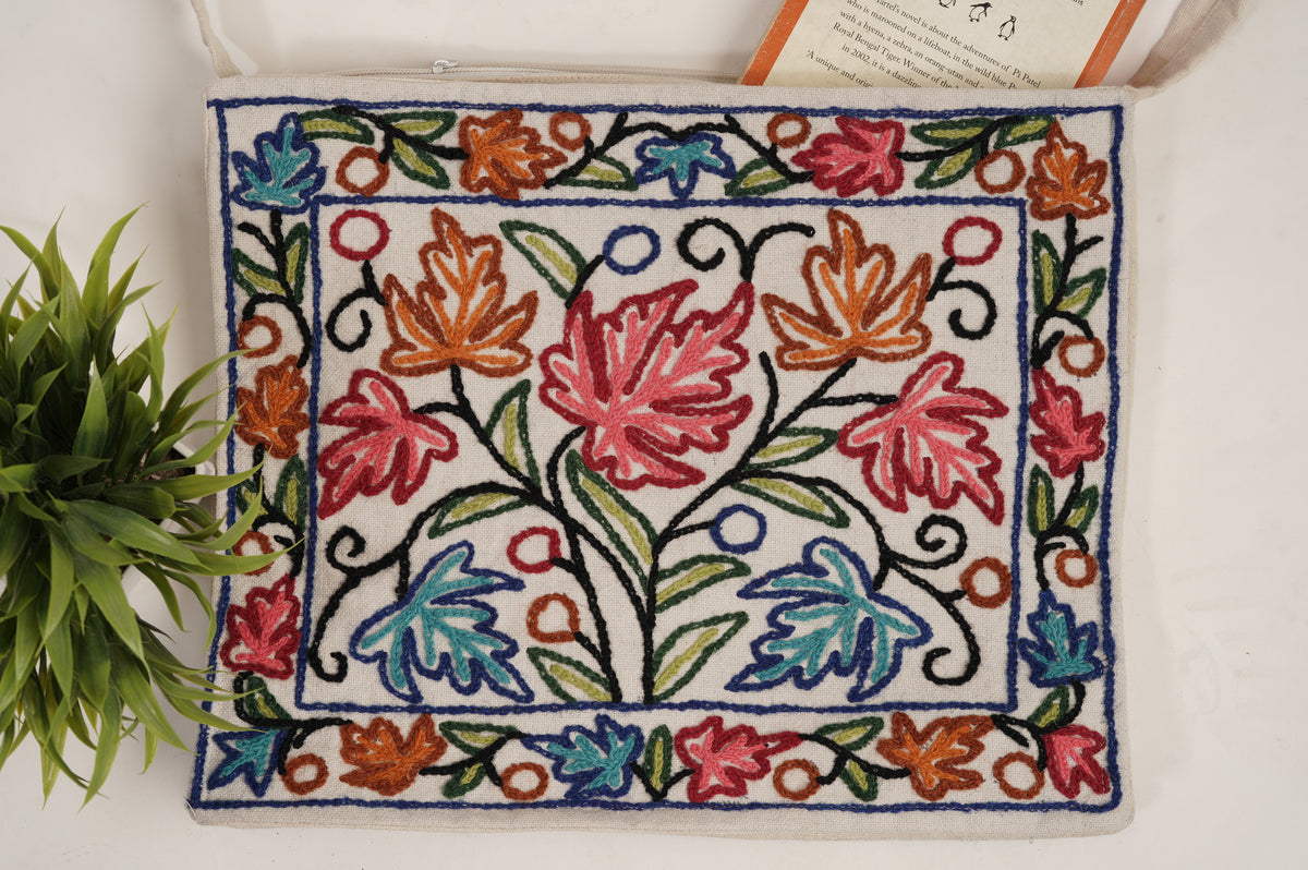 Cream cotton shoulder bag laid flat, with red, blue and orange Aari leaf motifs, green stems and navy outlines