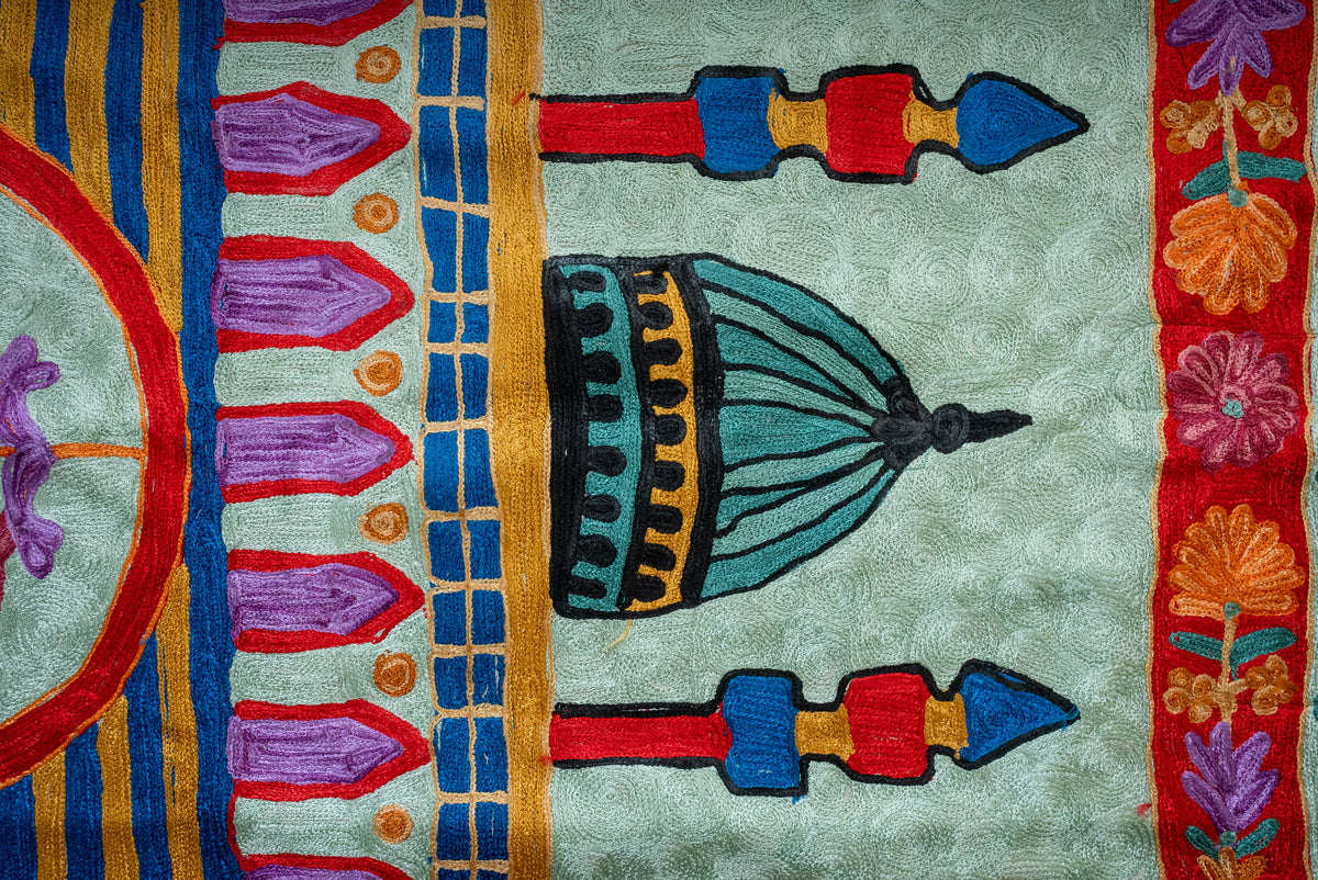 Detail of a green dome between red, blue and gold minarets above purple arches on the prayer mat