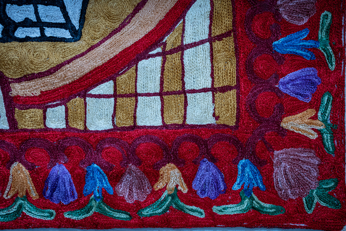 Close-up of blue, purple and cream embroidered flowers with green leaves along the prayer mat's red border