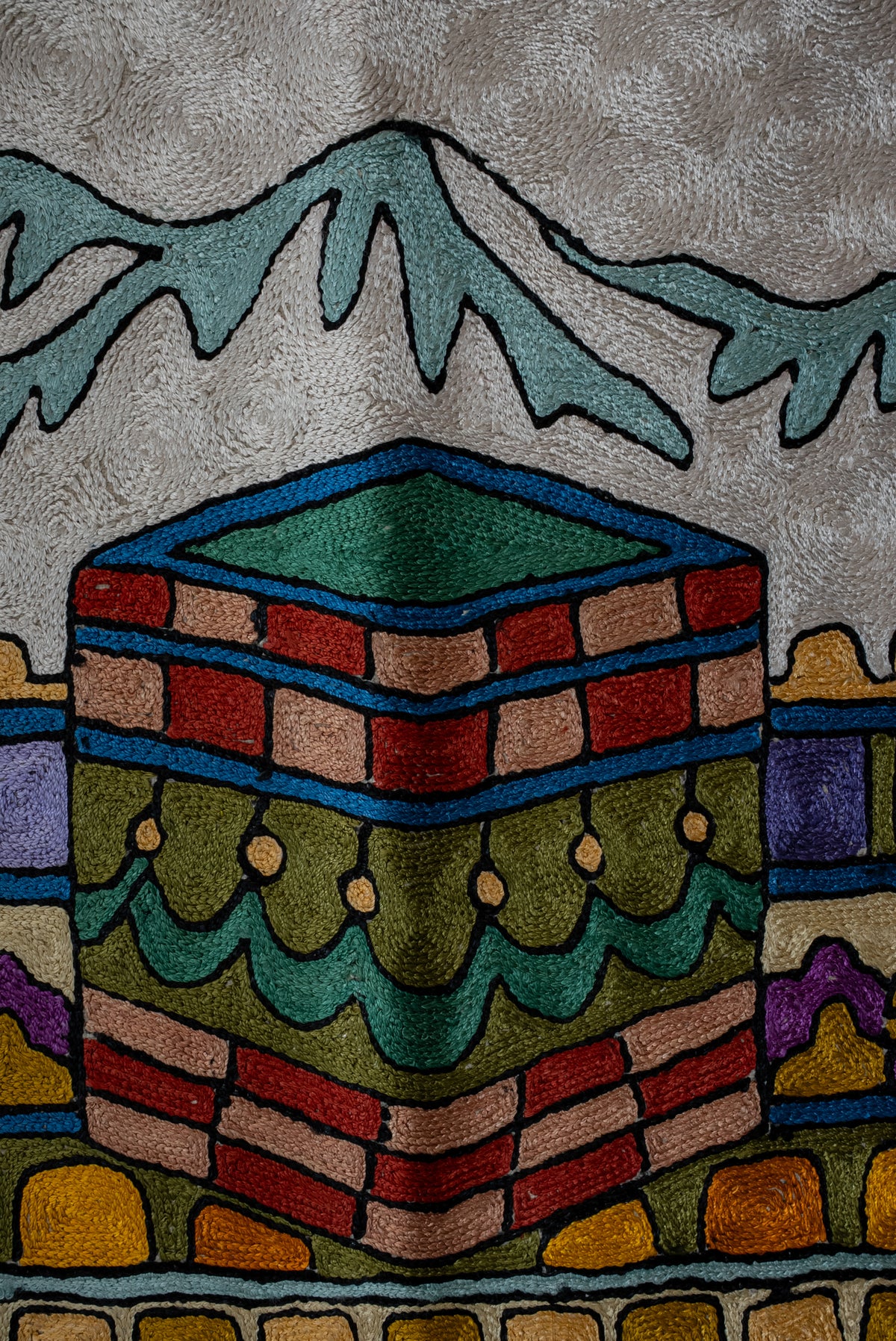 Detailed view of the embroidered Kaaba motif with red and pink blocks, a green top and pale blue mountains behind