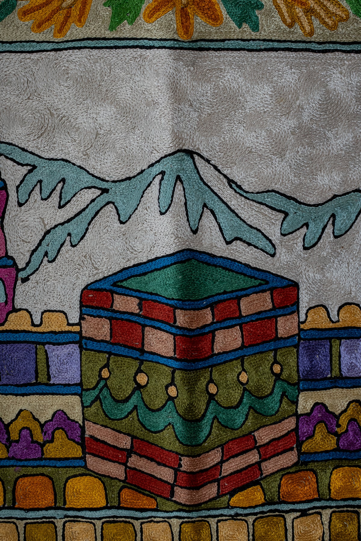 Close-up of the prayer mat's colourful Kaaba motif beneath pale blue mountains