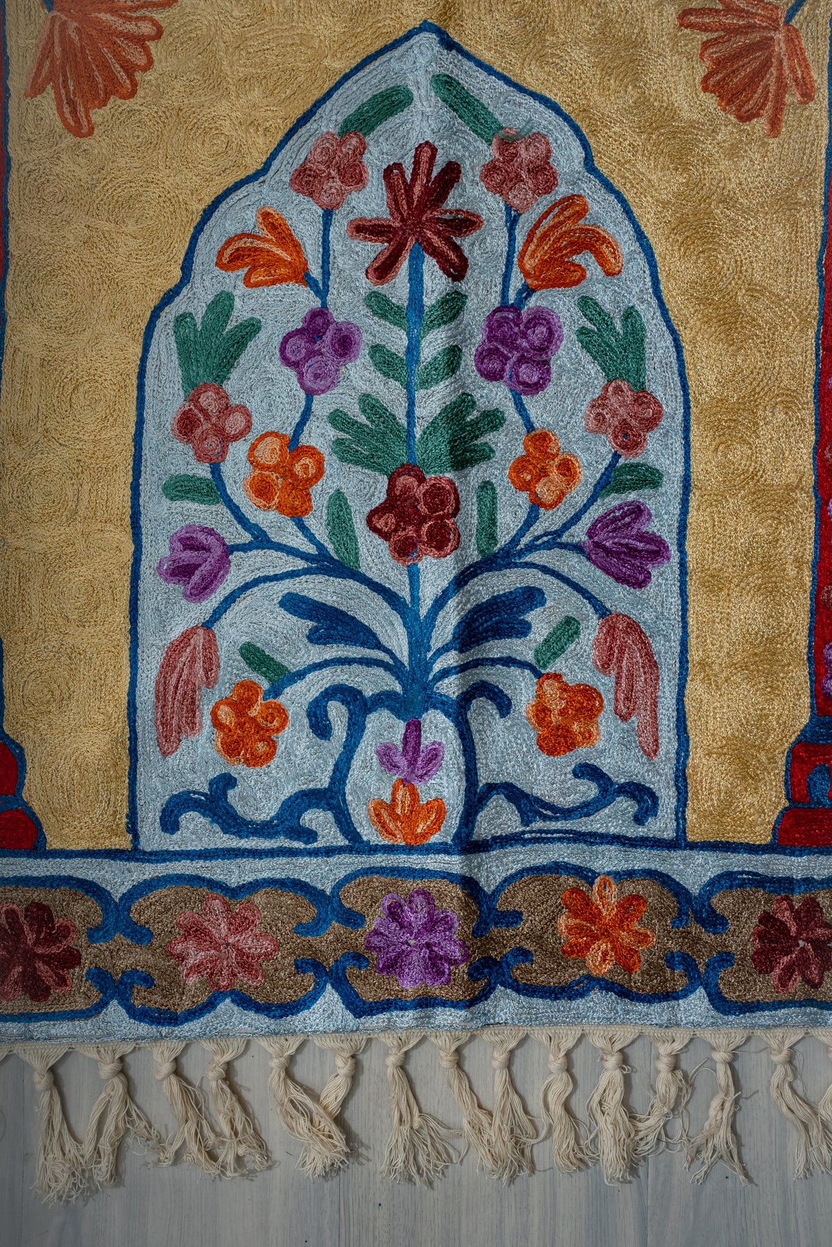 Lower end of the prayer mat with a multicoloured floral tree inside a pale blue arch and cream tassels